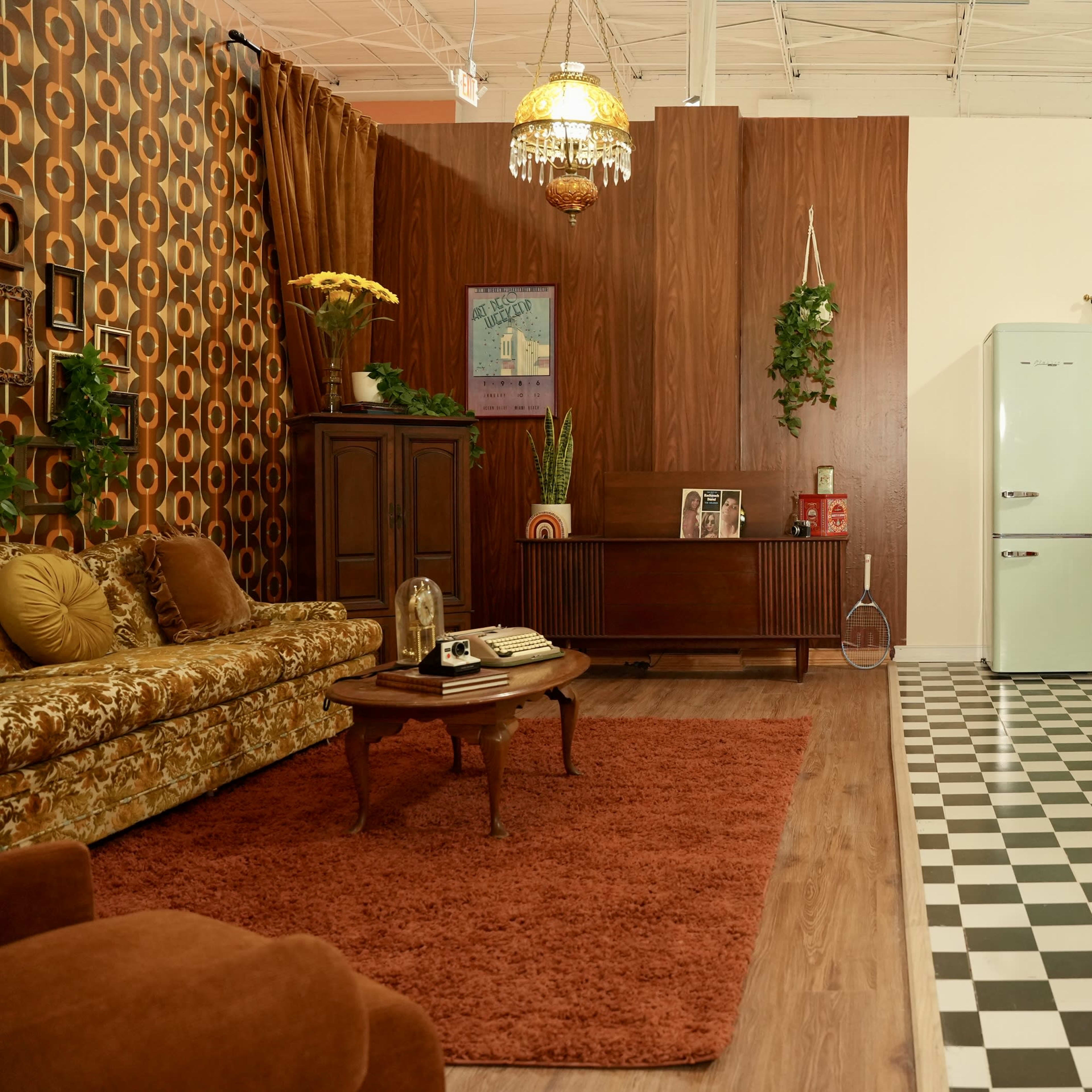 A retro-style living room and kitchen setup, featuring dark wood paneling, a patterned wall, vintage furniture, and a checkered floor.