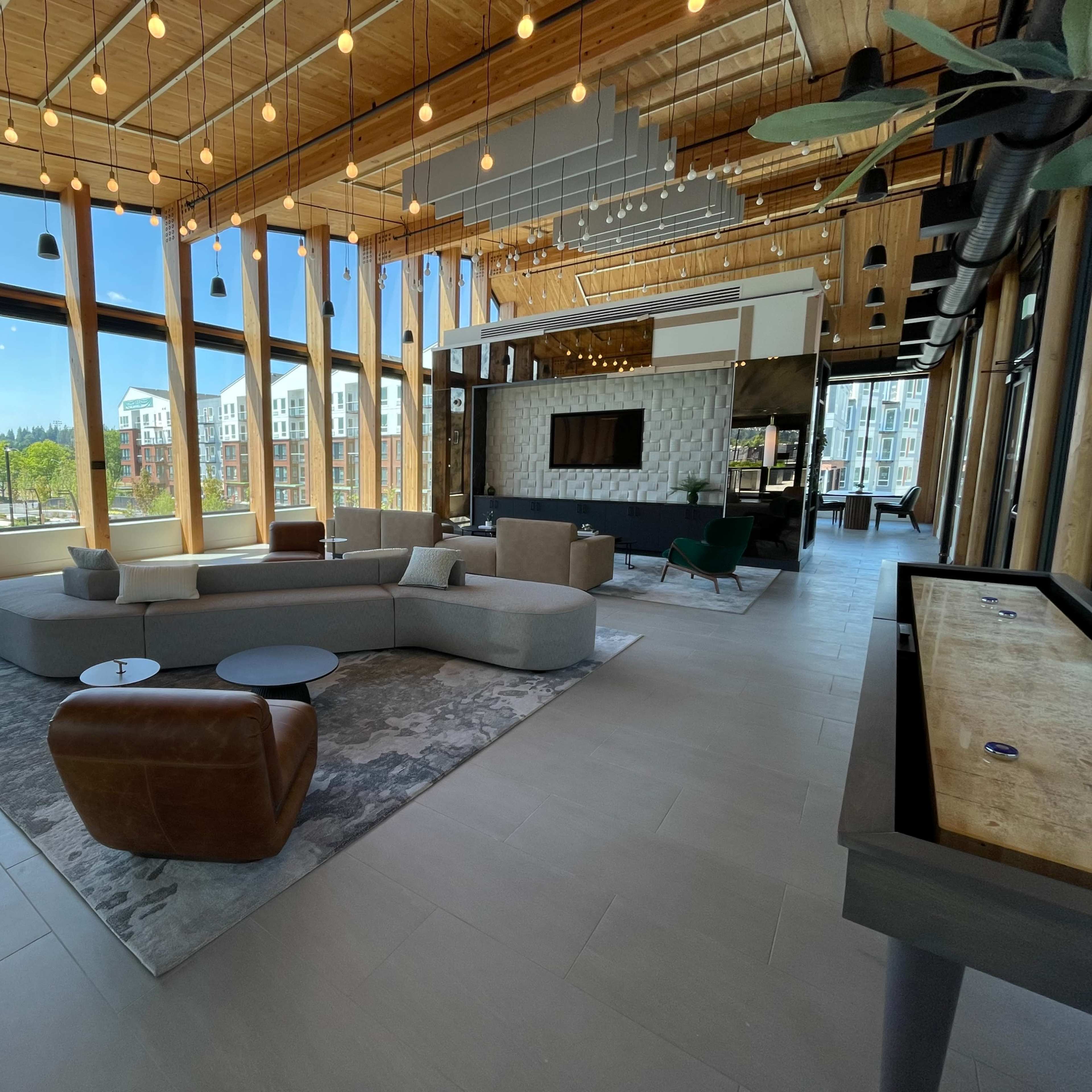 A spacious, modern lounge area with large windows, wooden beams, and a combination of sofas and chairs arranged around a central coffee table.