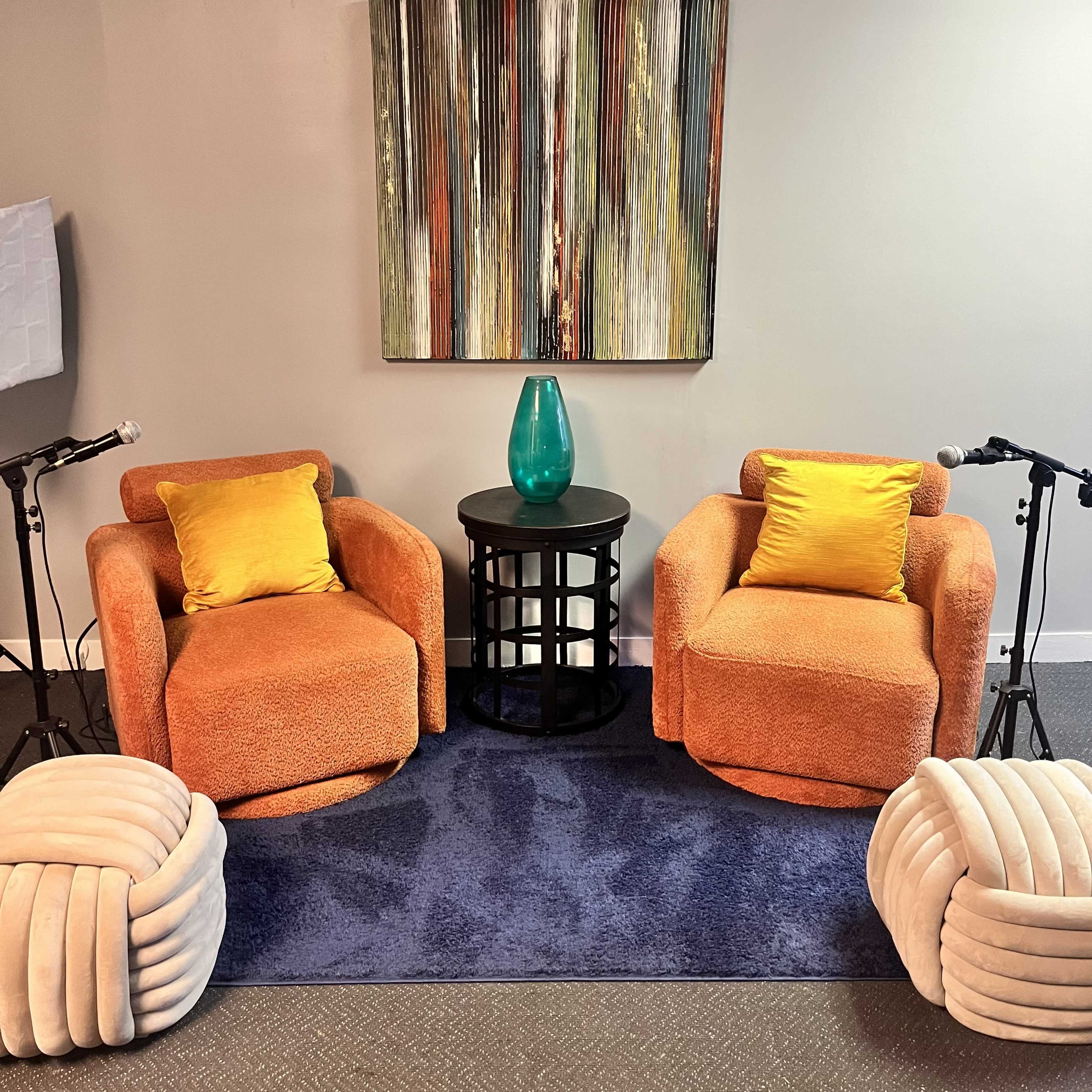 A cozy seating area with two orange armchairs, a blue rug underneath, a round black table with a teal vase, and two microphones positioned nearby.