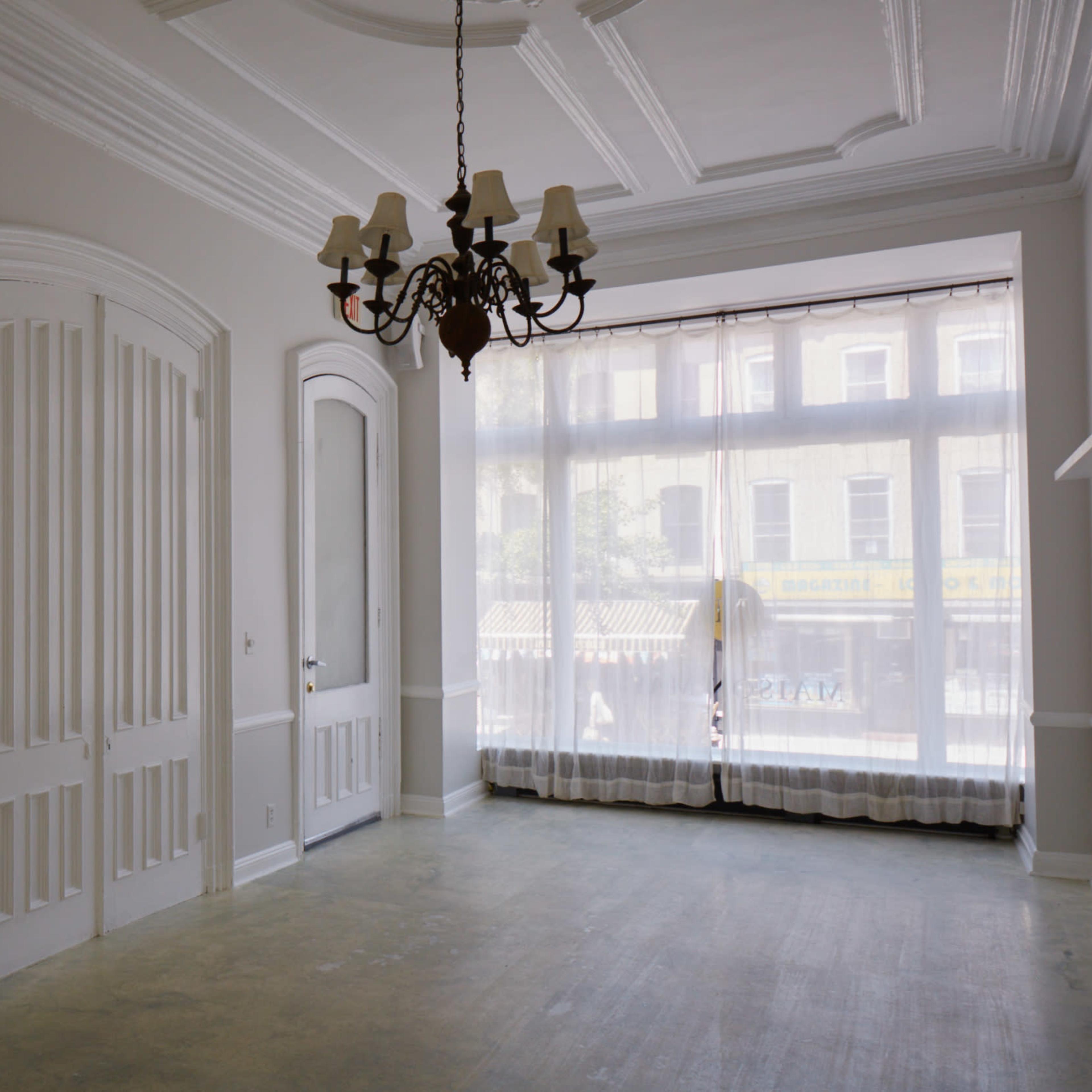 A spacious, empty room with white walls, a chandelier, and large windows dressed with sheer curtains.