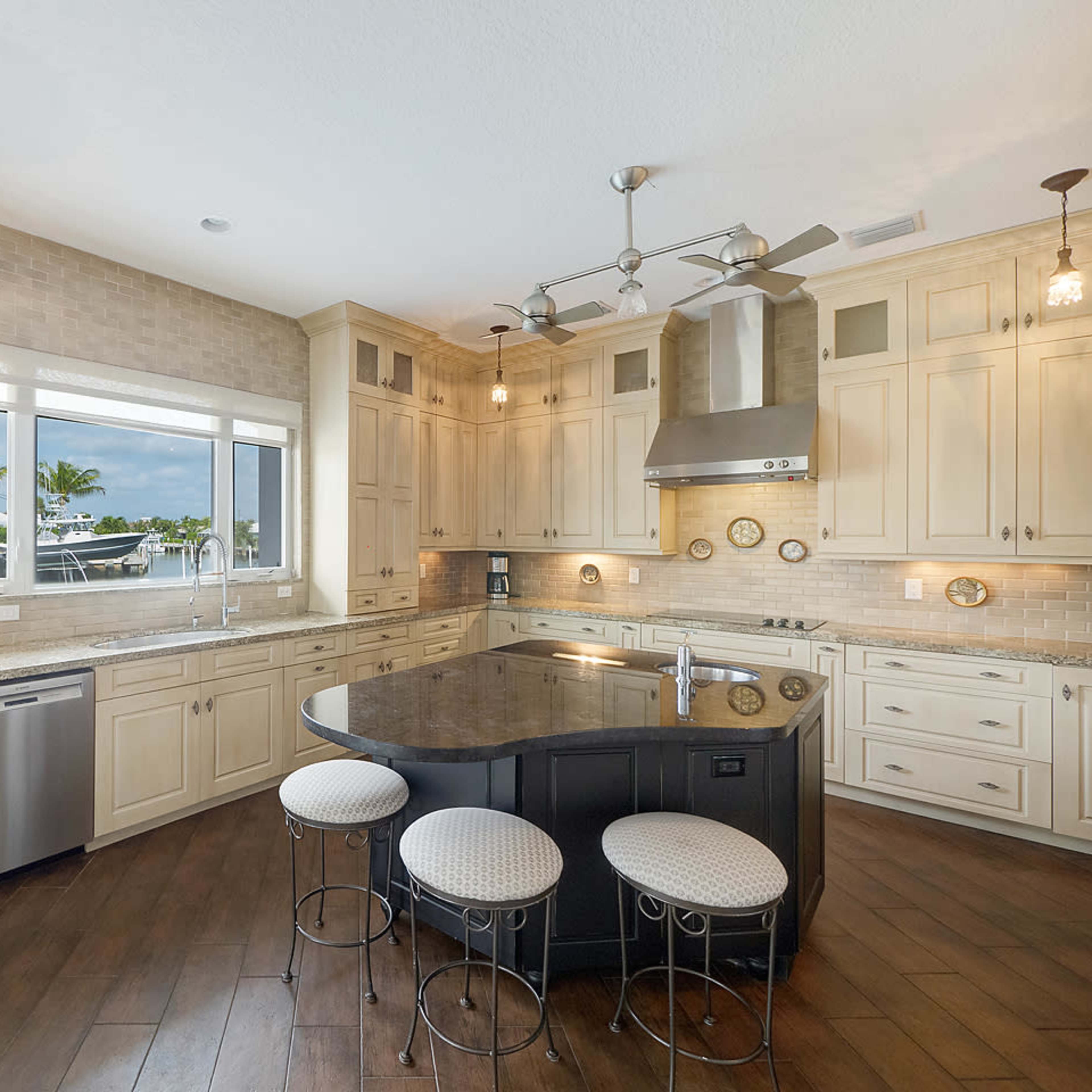 A modern kitchen features cream-colored cabinets, a large dark island, stainless steel appliances, and a window overlooking a boat dock.