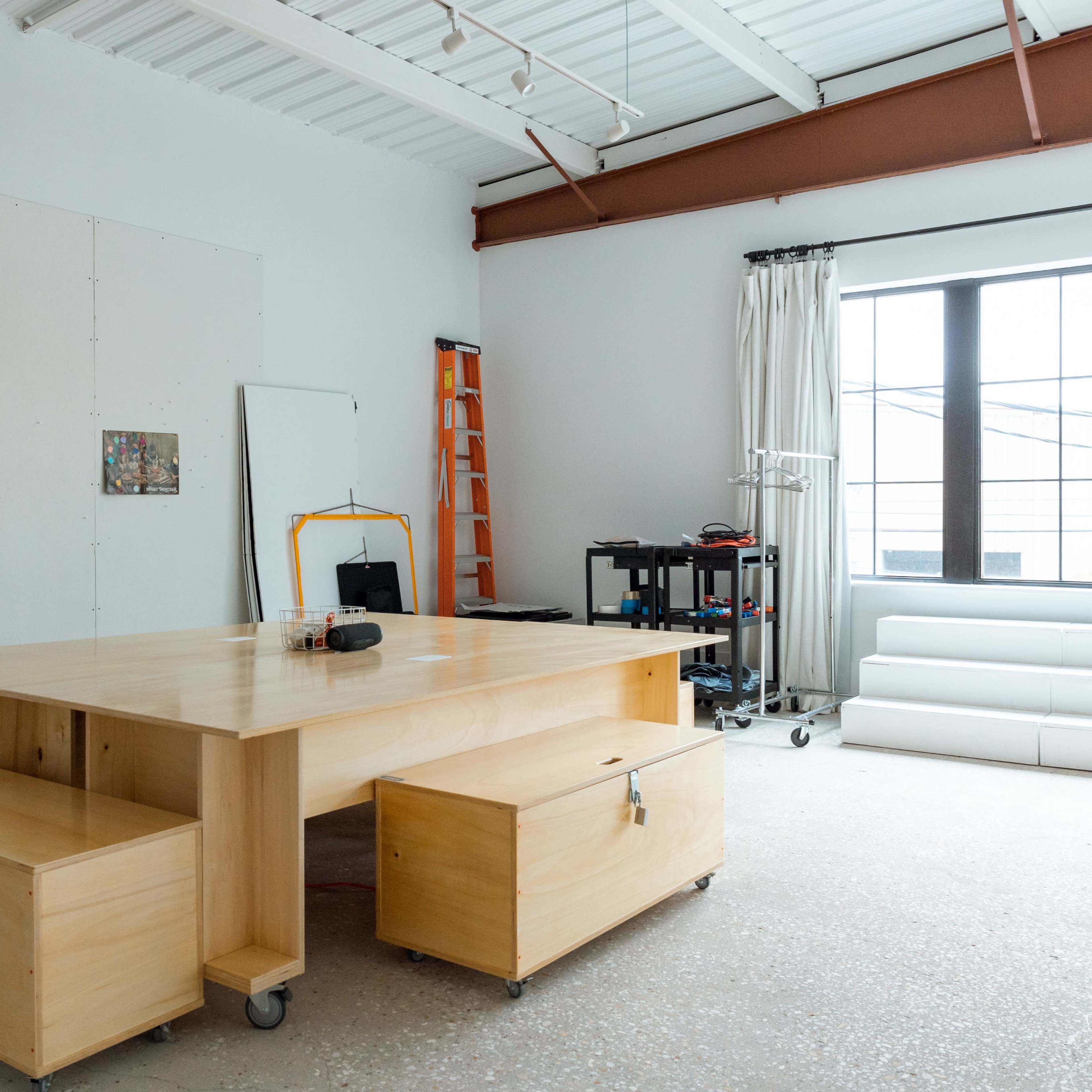 A spacious studio with a wooden table on wheels, two drawers, and various items, including a ladder and artwork pinned to the wall.