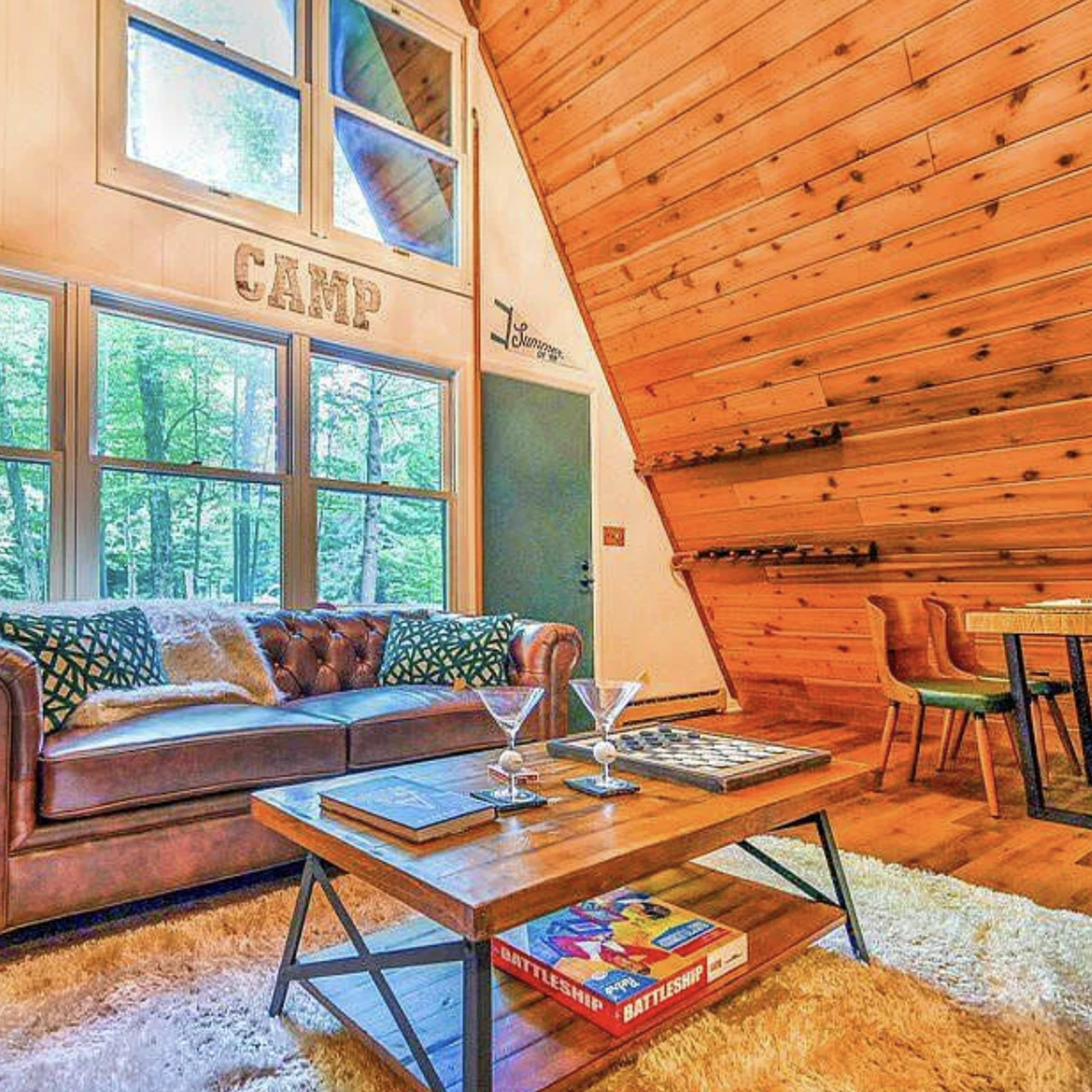 A cozy living room with a leather sofa, wooden walls, and large windows offering views of trees outside.