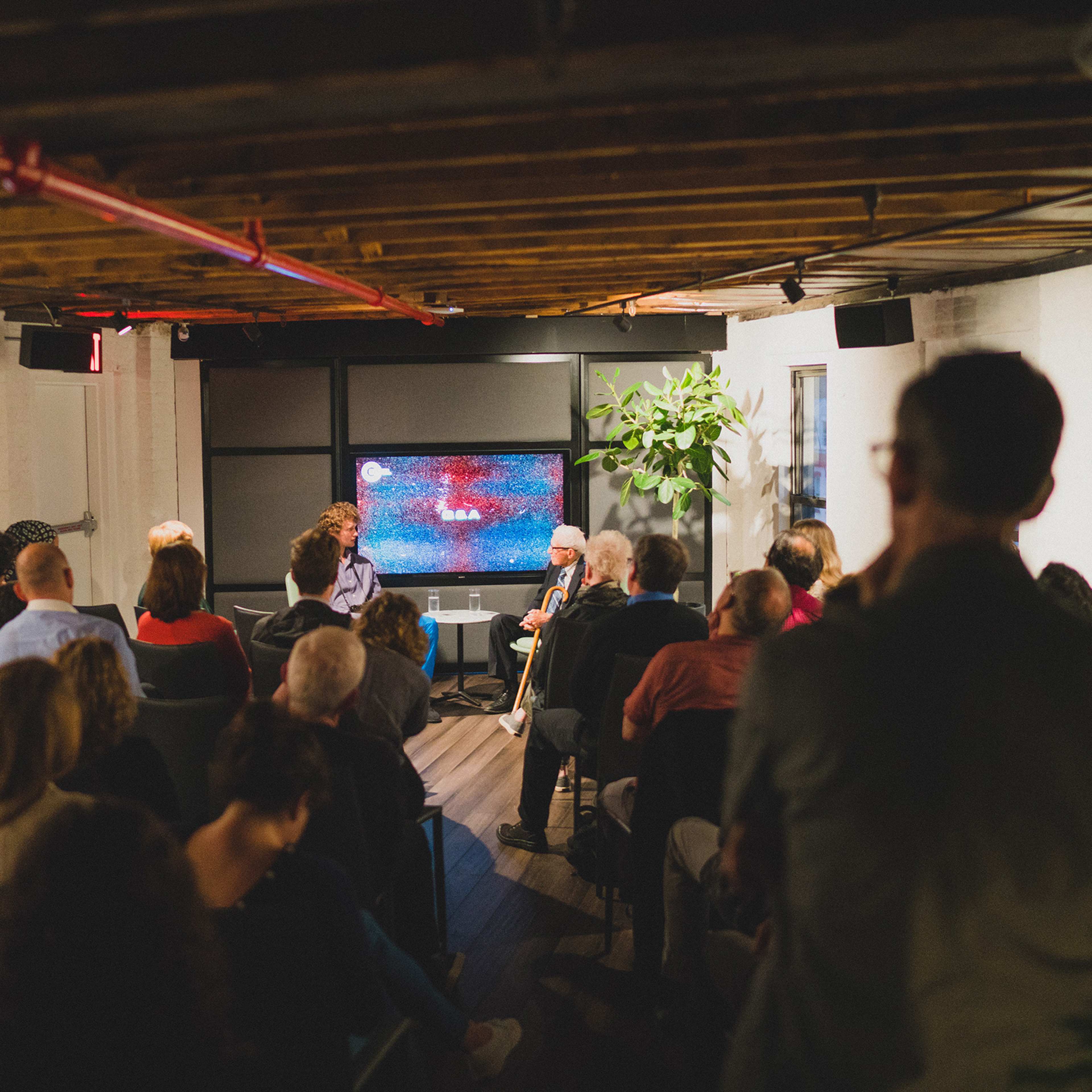 A crowd of people sits in an indoor venue with exposed wooden beams, watching a presentation featuring two speakers in front of a screen displaying colorful visuals.