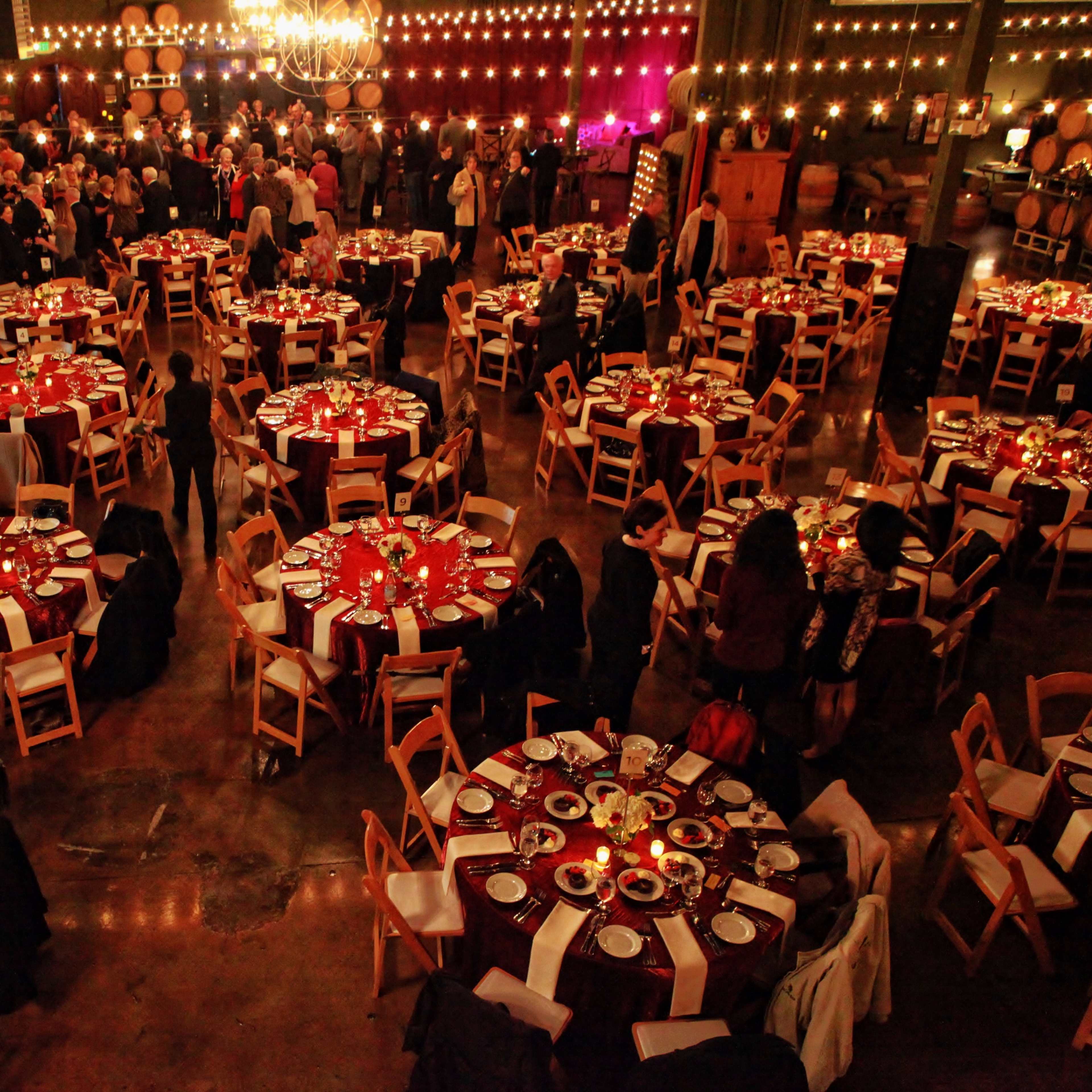 A large banquet hall filled with people gathered around elegantly set round tables, with decorations and string lights in the background.