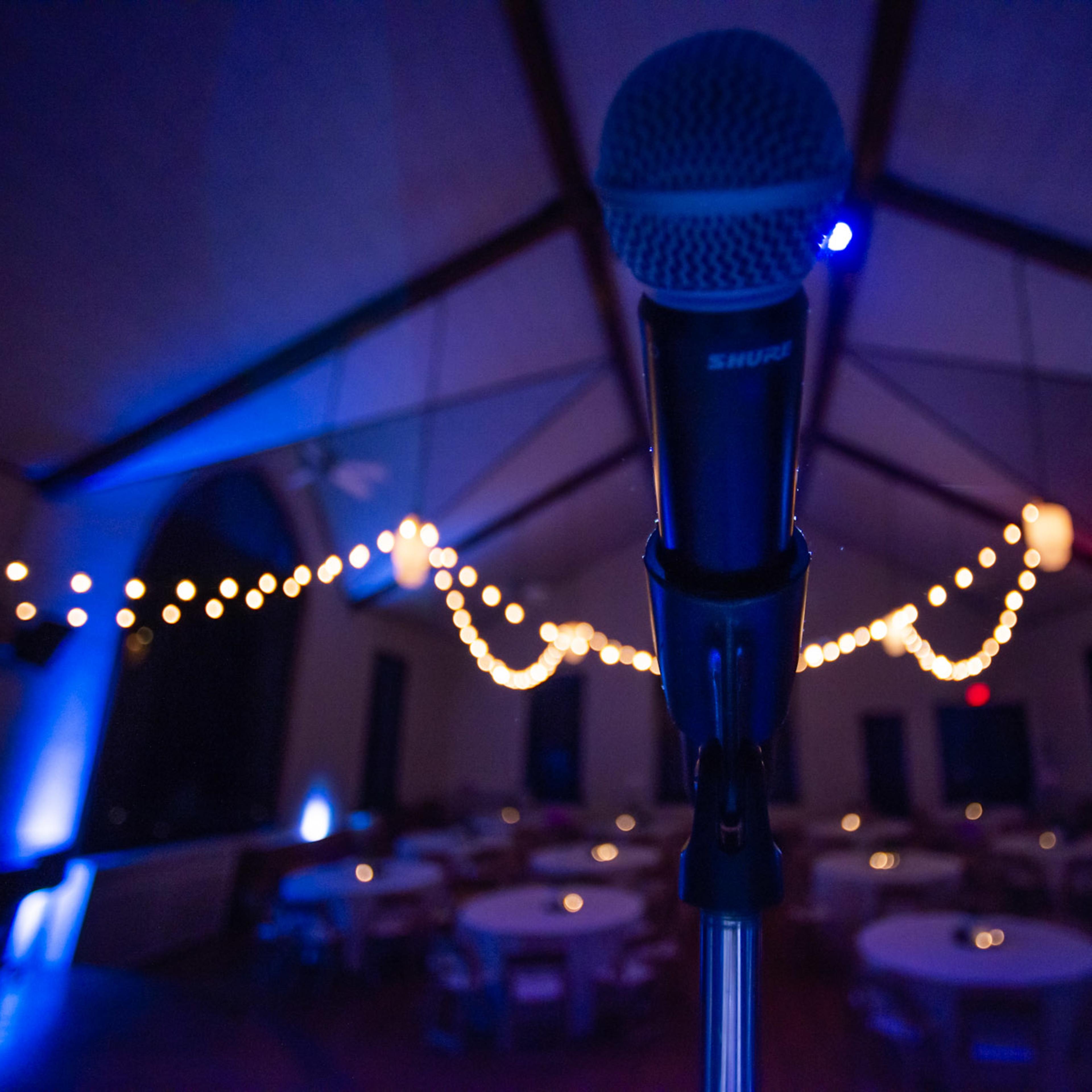 A microphone stands in the foreground with a decorated venue featuring tables and string lights in the background.