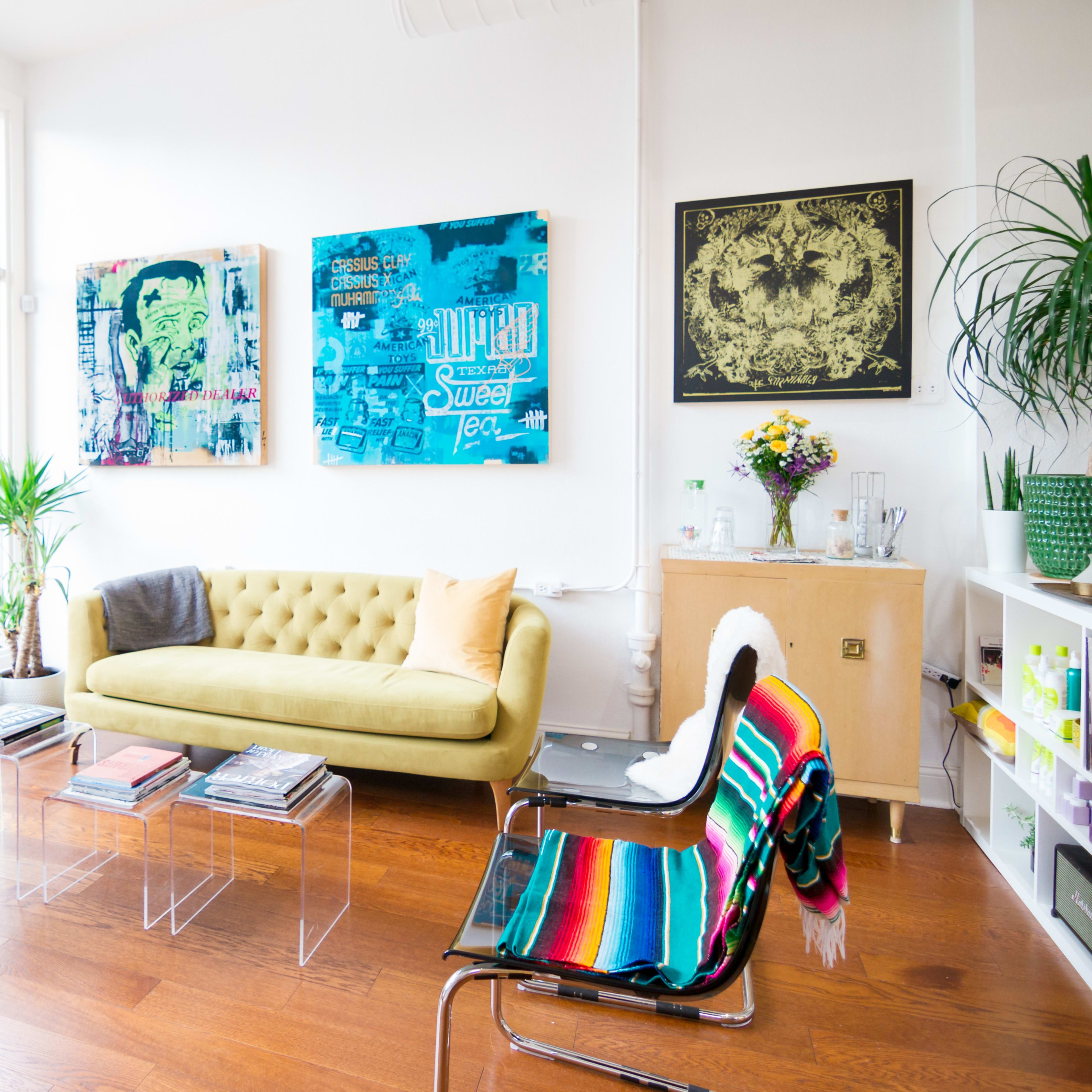 A brightly lit living room with a yellow tufted sofa, colorful artwork on the walls, and a mix of modern furniture, including a clear coffee table and a striped chair.