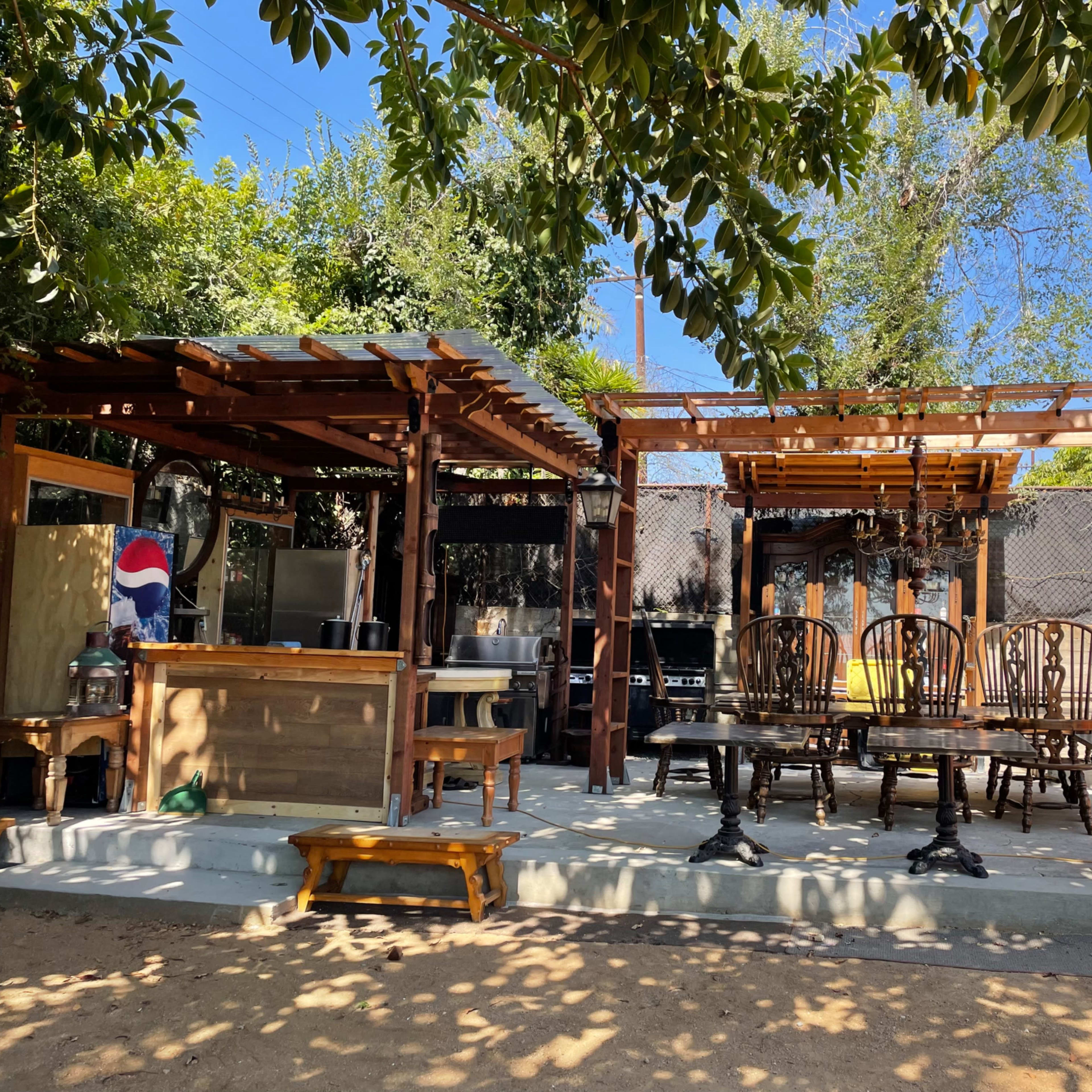 An outdoor seating area with wooden furniture and a bar structure, surrounded by trees and sunlight.