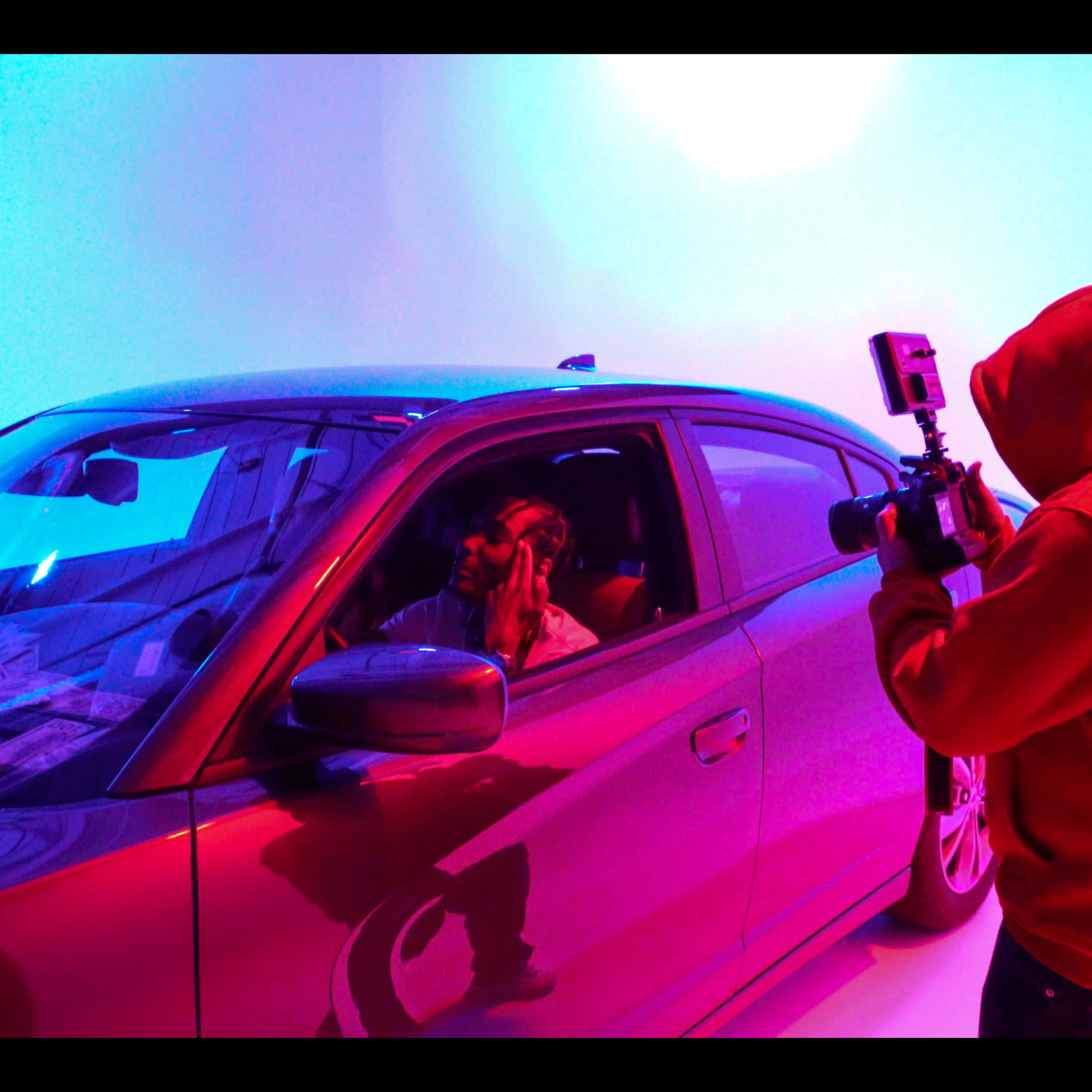 A person wearing a red hoodie photographs an individual inside a car against a colorful illuminated backdrop.