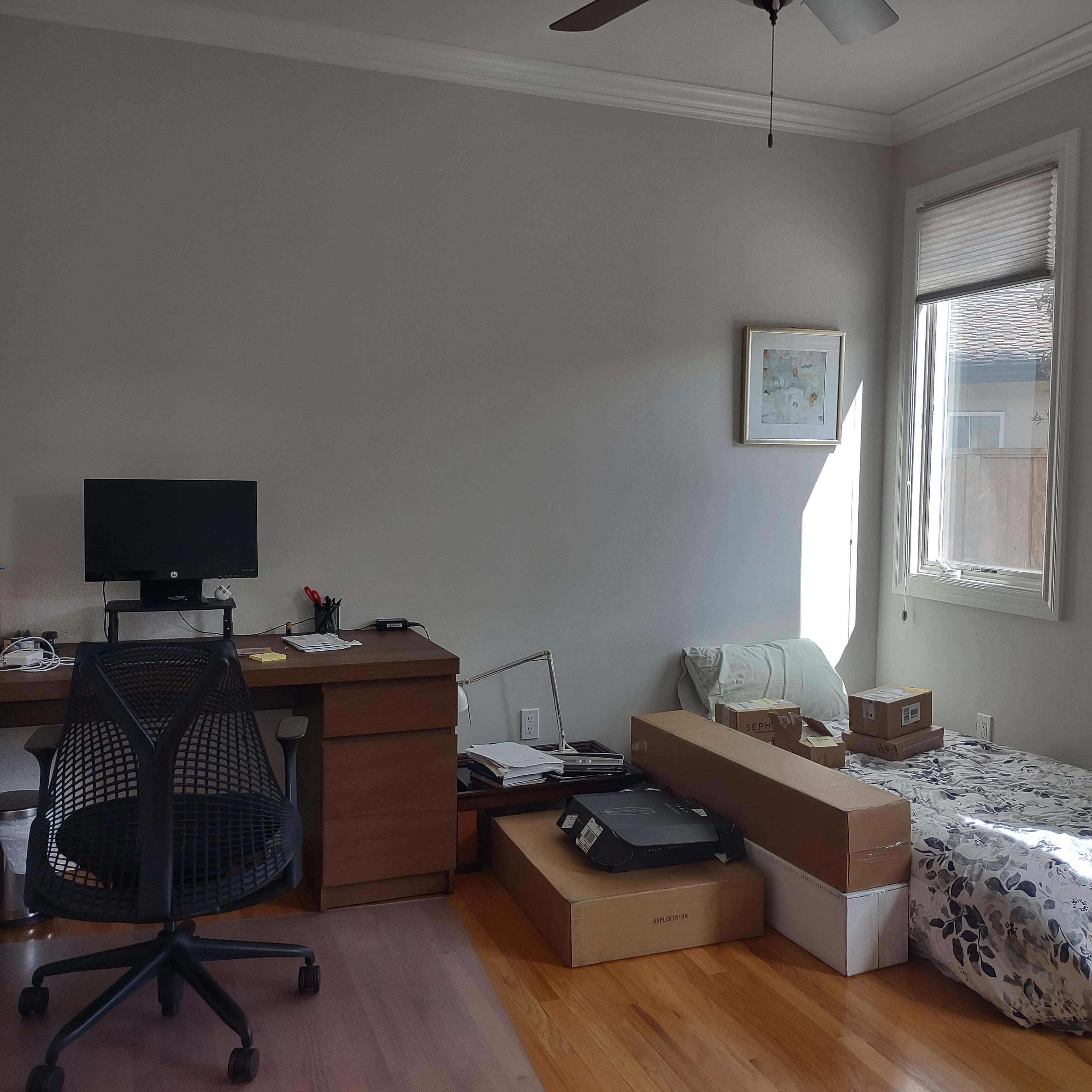 A minimalist office space features a desk with a computer and office supplies, a chair, and a bed surrounded by boxes and a small window letting in natural light.