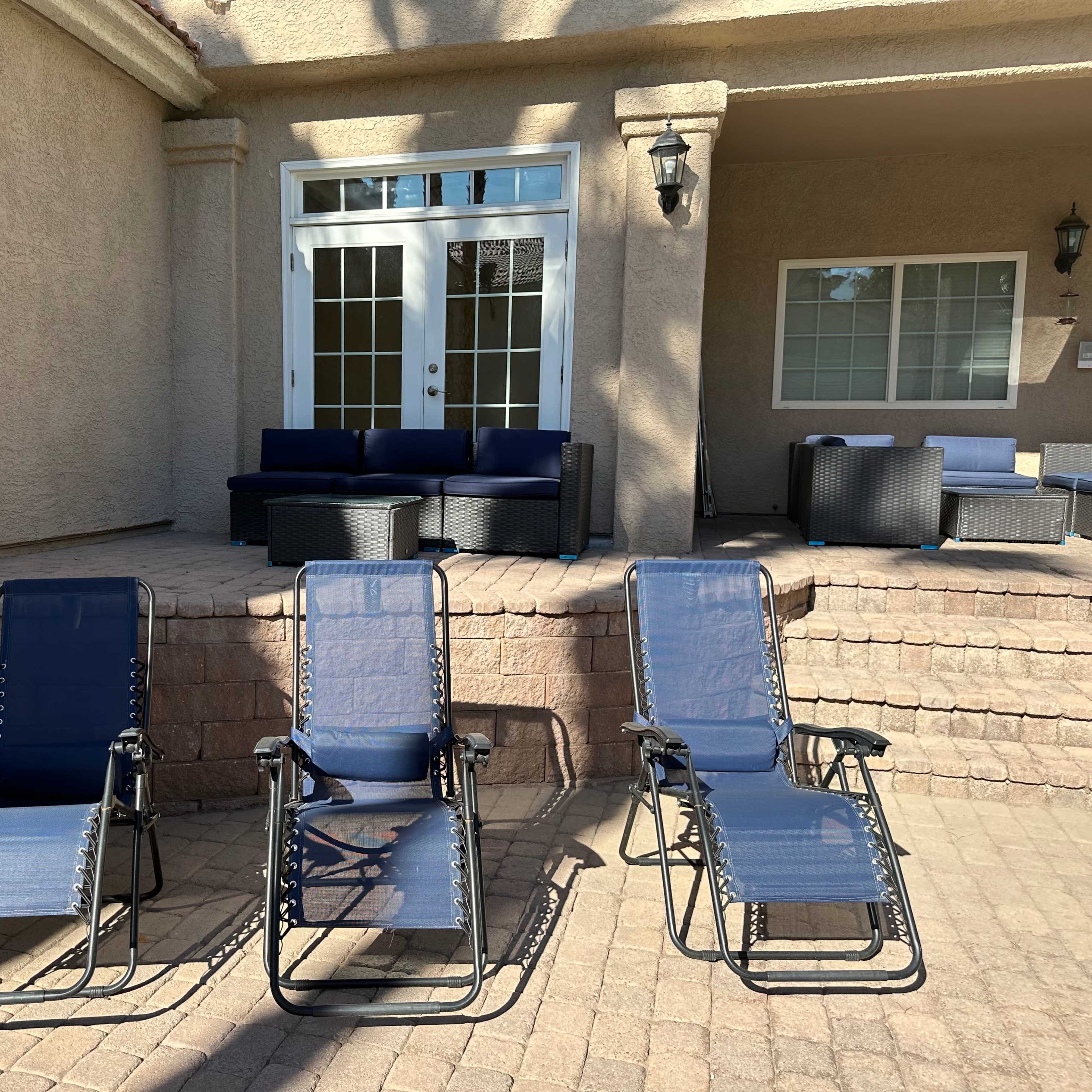 Three reclining chairs are positioned on a stone patio in front of a house with a seating area visible through large glass doors.