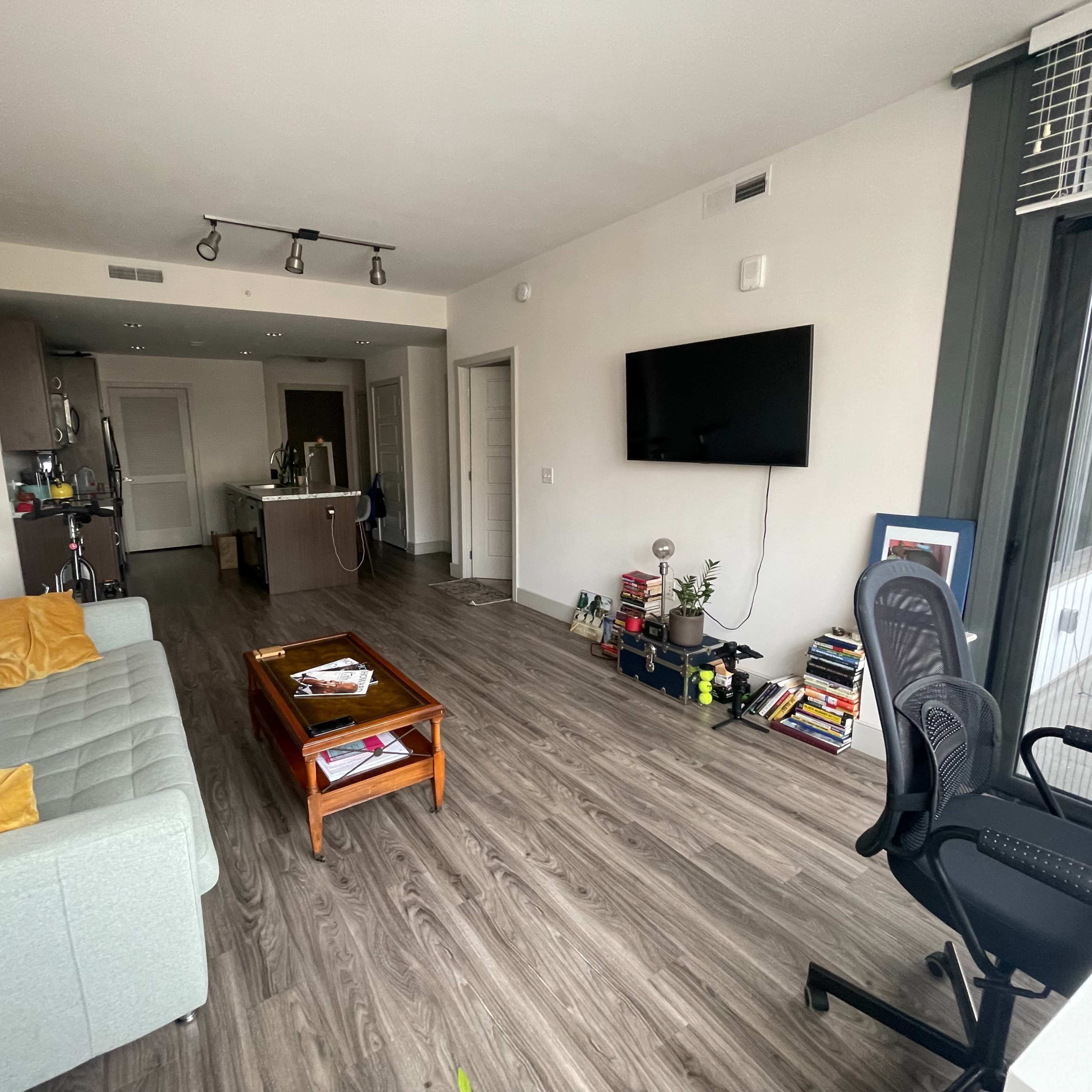A modern, light-filled apartment features a gray sofa, a wooden coffee table, and a large wall-mounted television, with an open kitchen area visible in the background.