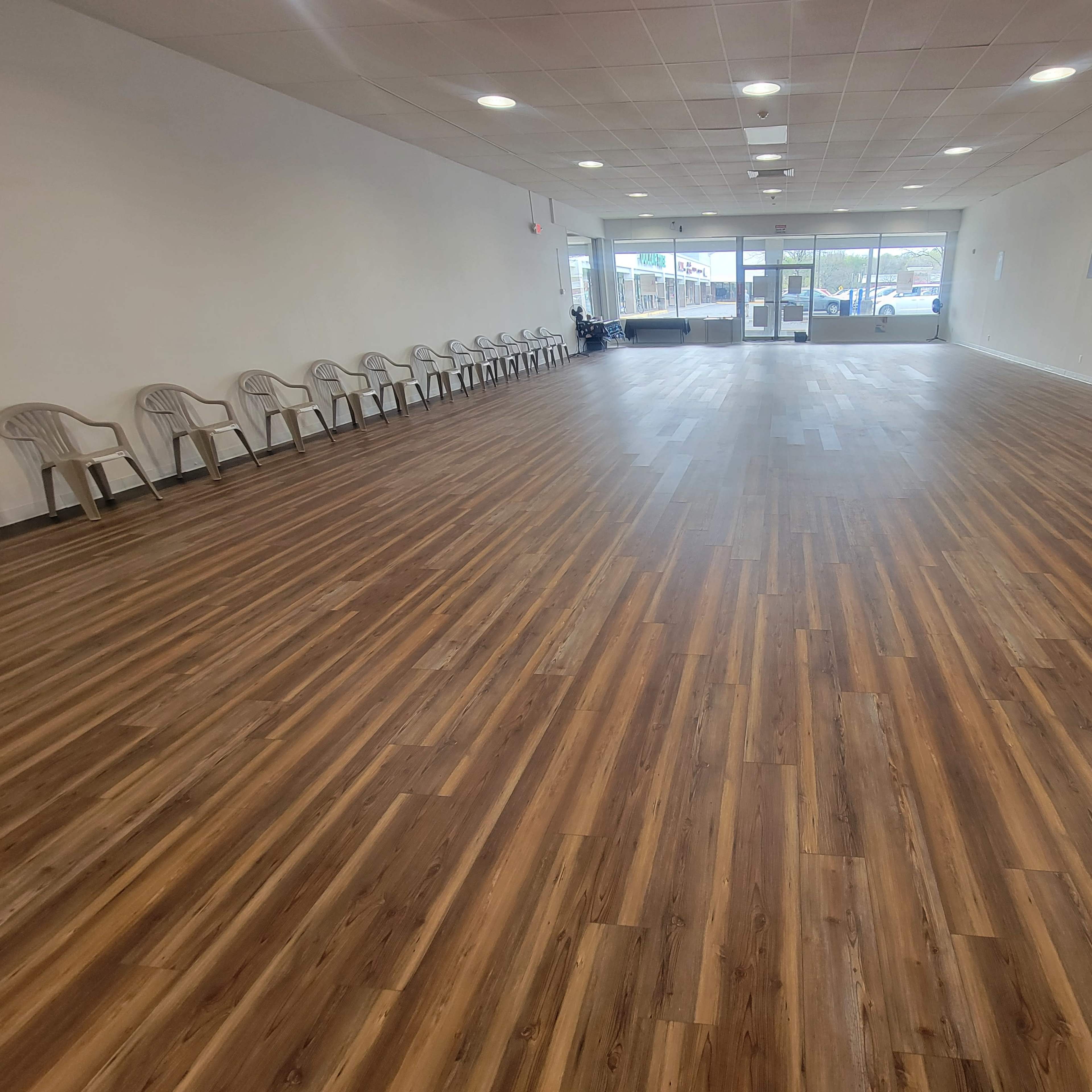 An empty room with a polished wooden floor and a row of plastic chairs along one wall, featuring large windows at the entrance.
