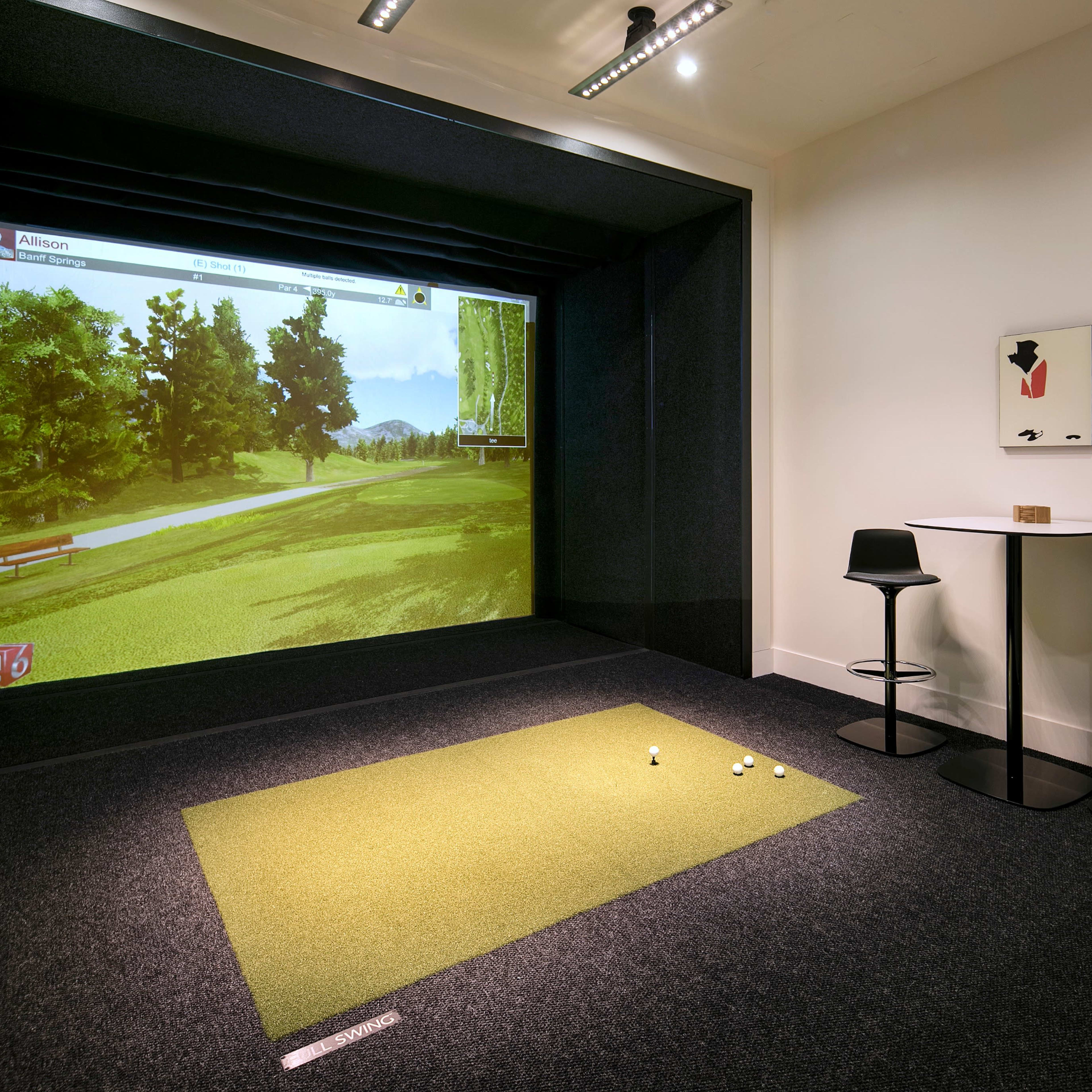 An indoor golf simulator setup with a large screen displaying a virtual golf course, a putting area with several golf balls, and a small table and chairs nearby.