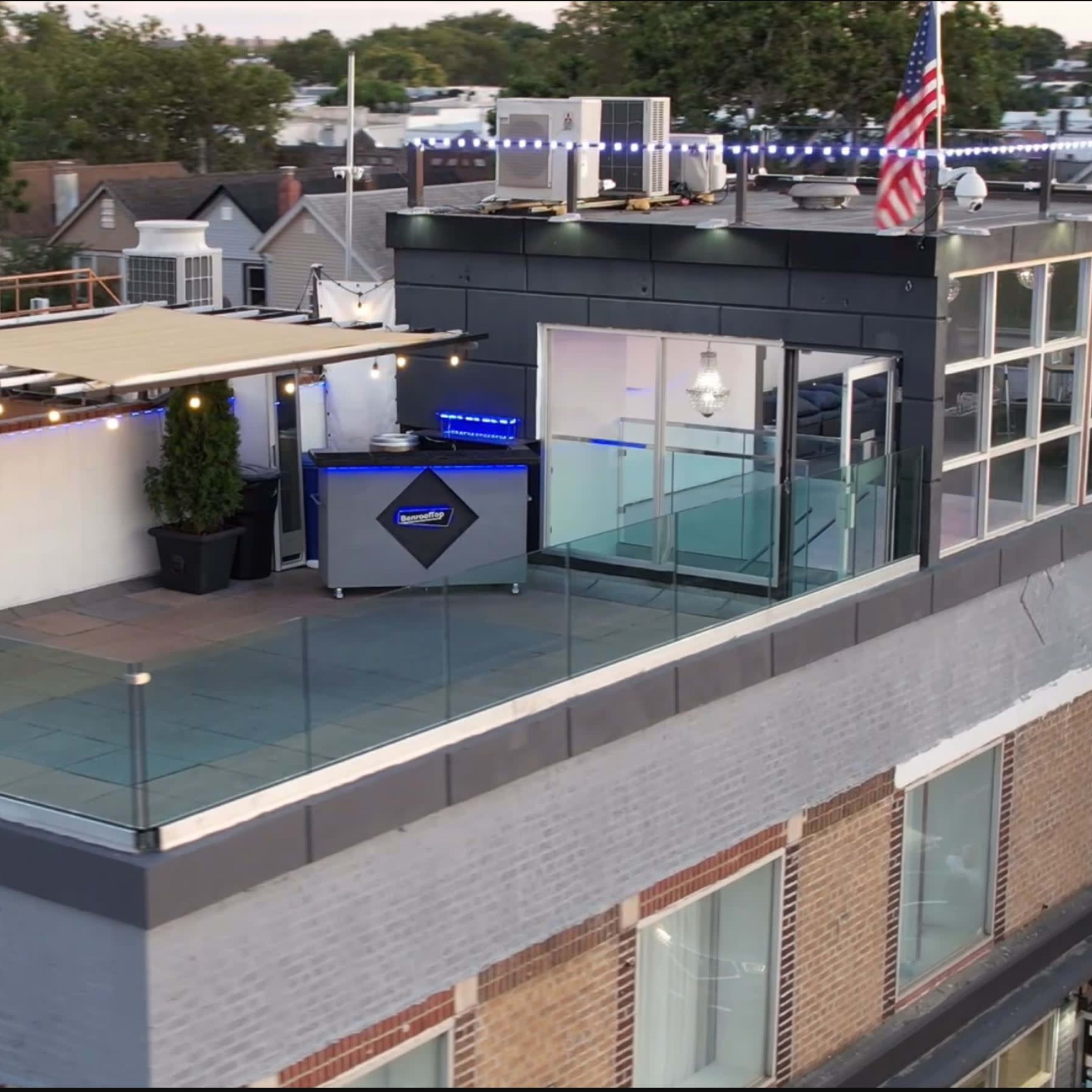 A rooftop terrace with glass railings, two potted plants, and a bar area, alongside an American flag and various structures in the background.