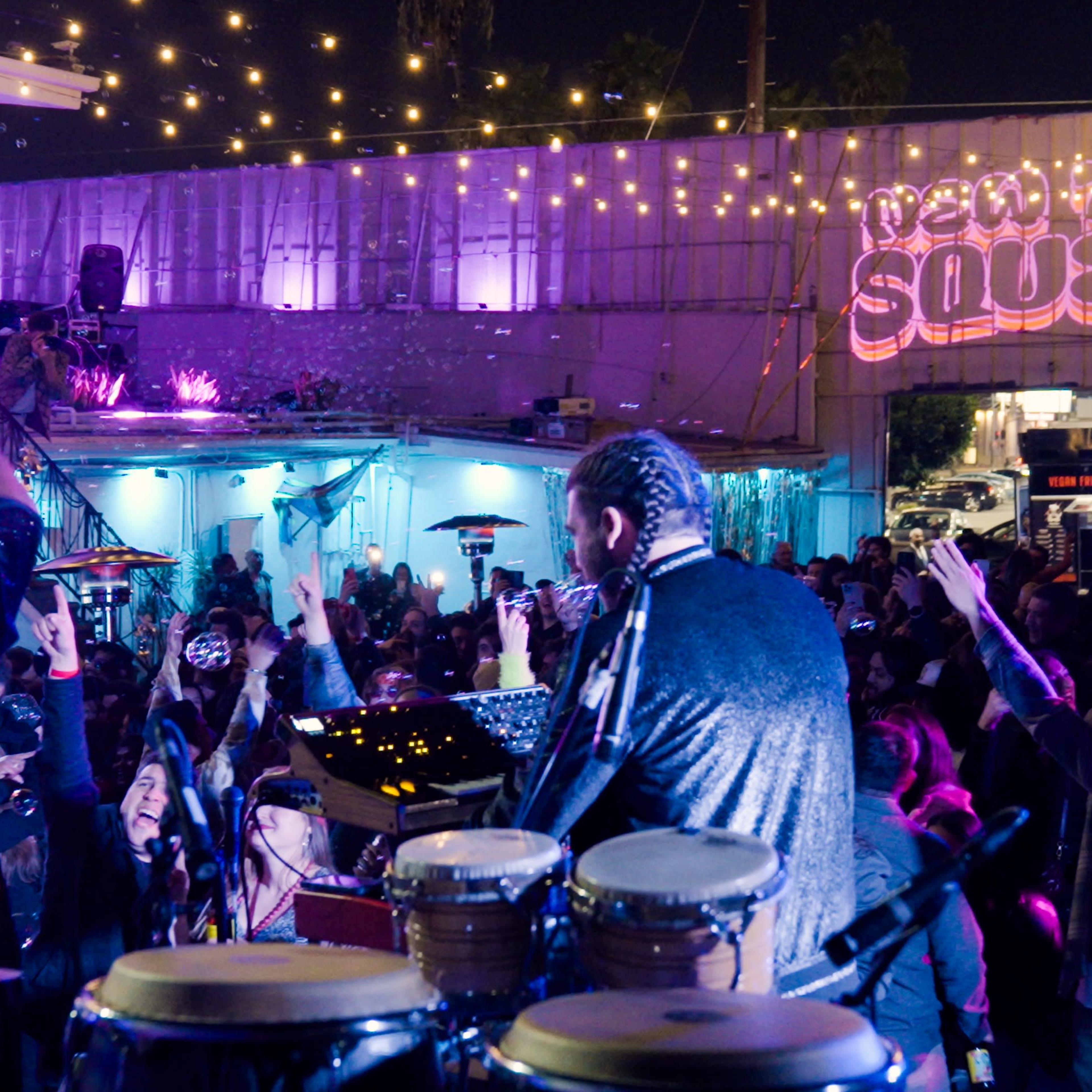 A lively outdoor party features a band performing on stage, surrounded by an energetic crowd and decorative string lights.