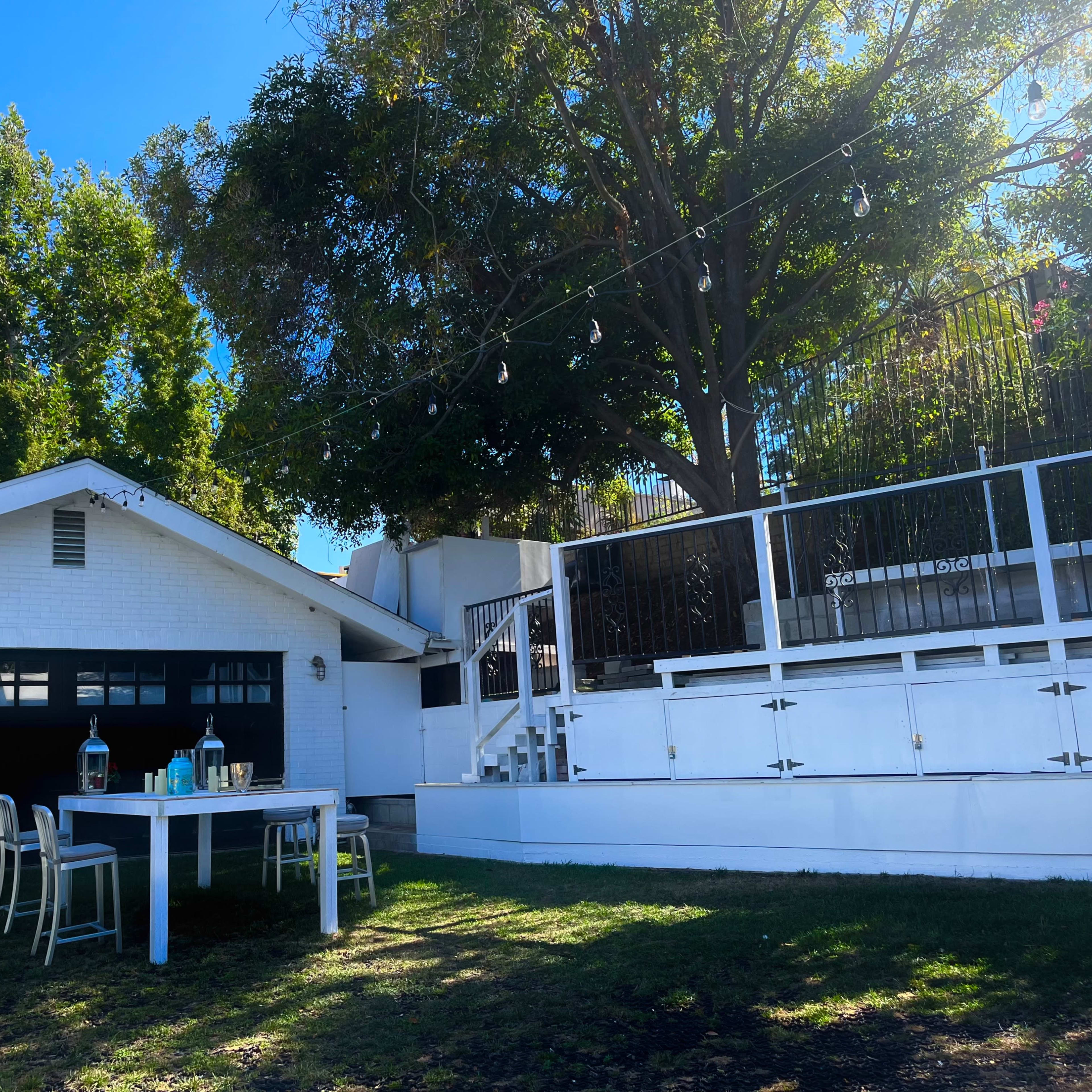 A white house with a garage sits beside a patio area featuring a table and chairs beneath a large tree.