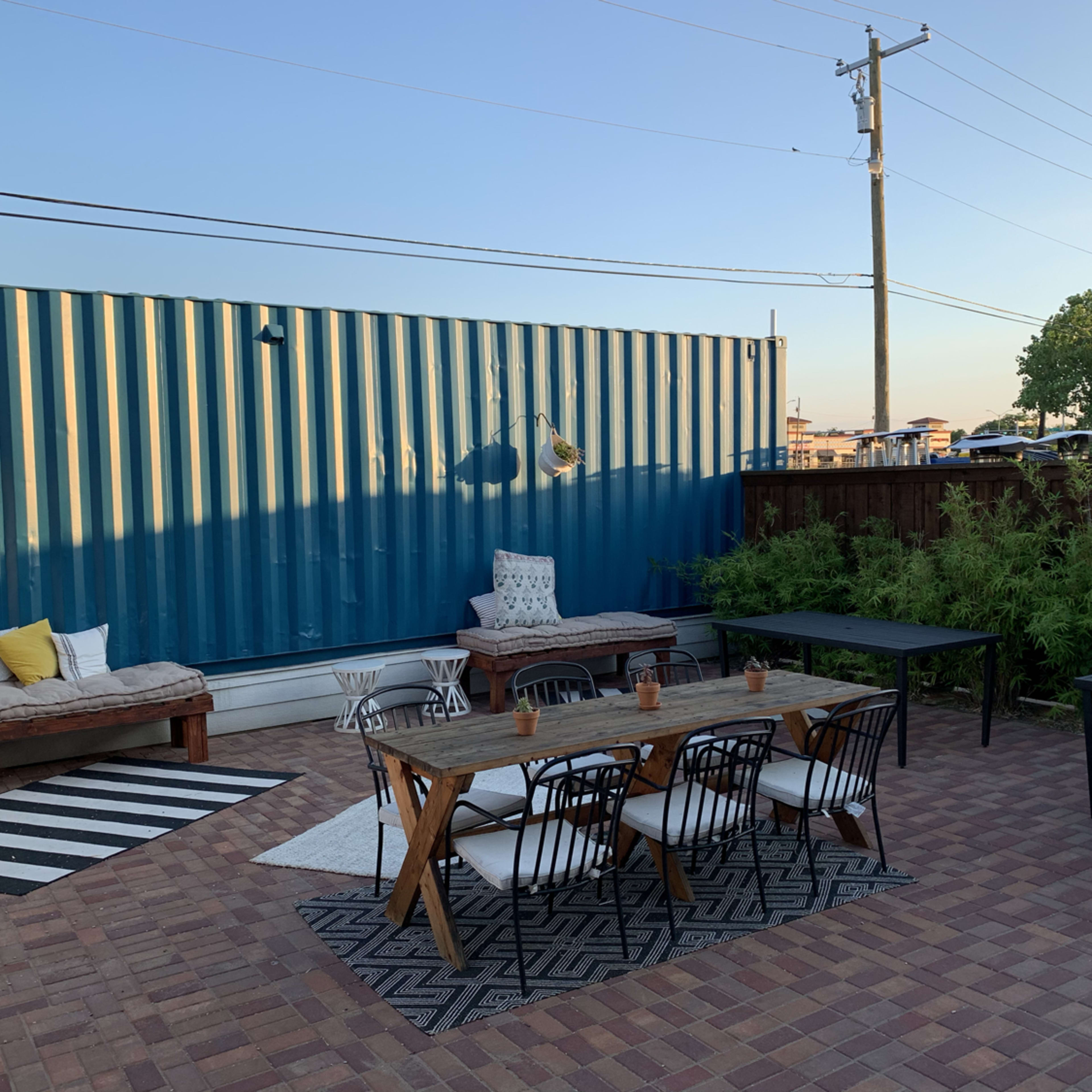 The scene features a patio area with a seating arrangement, a large blue shipping container in the background, and several outdoor tables on a brick surface.
