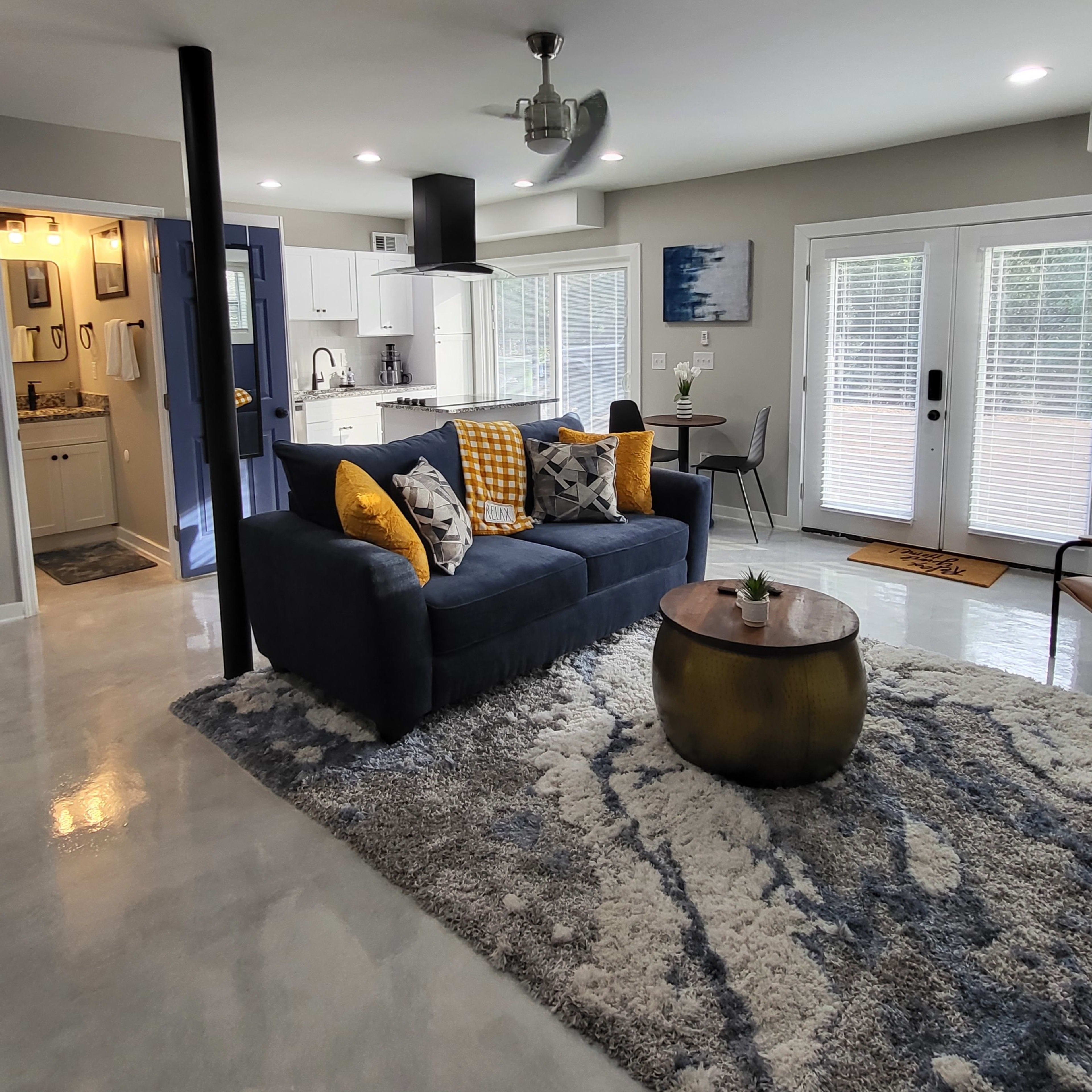A modern living room with a dark sofa, a round coffee table, and large windows, accompanied by a kitchen area in the background.
