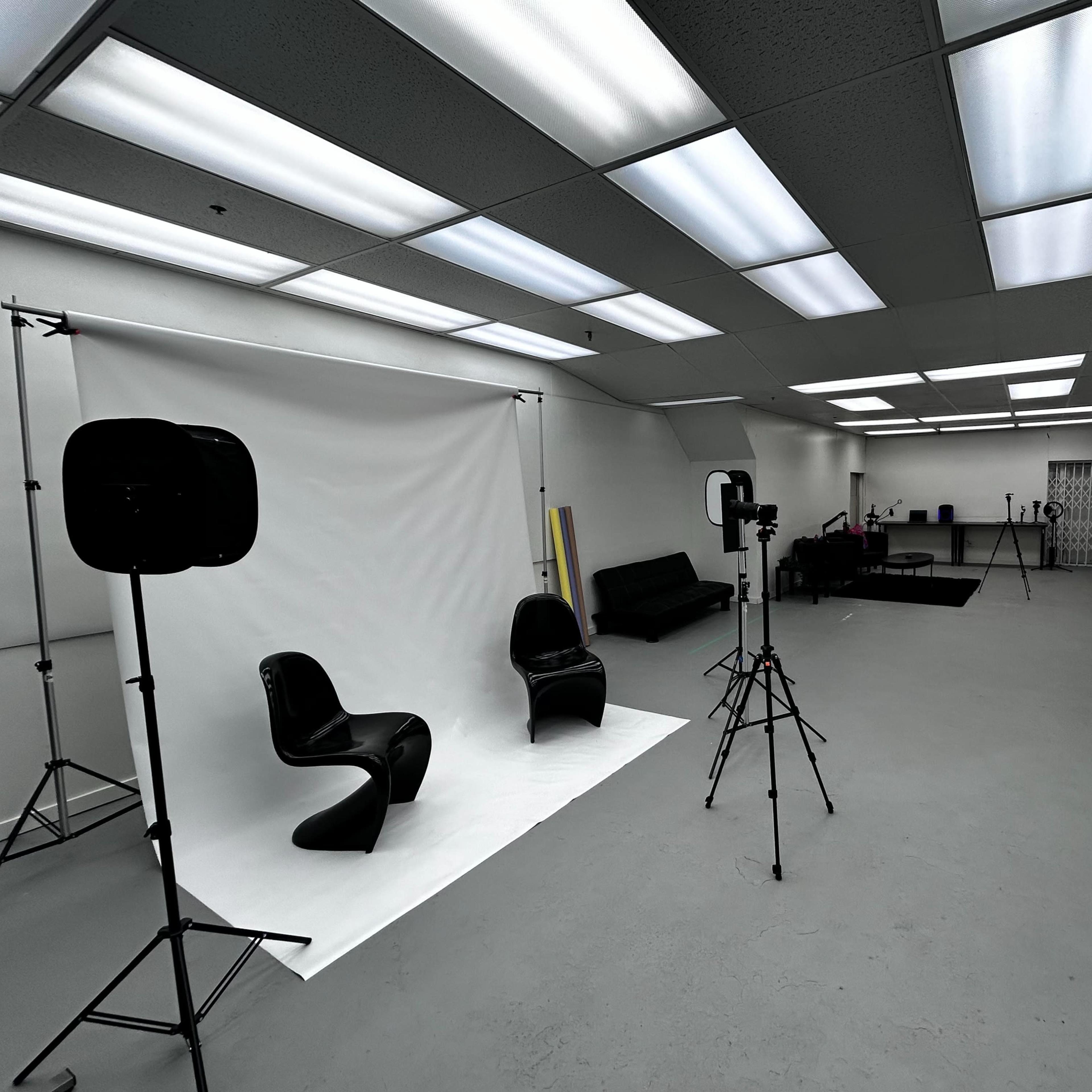 A photography studio with two black chairs positioned in front of a white backdrop, surrounded by lighting equipment and various props.