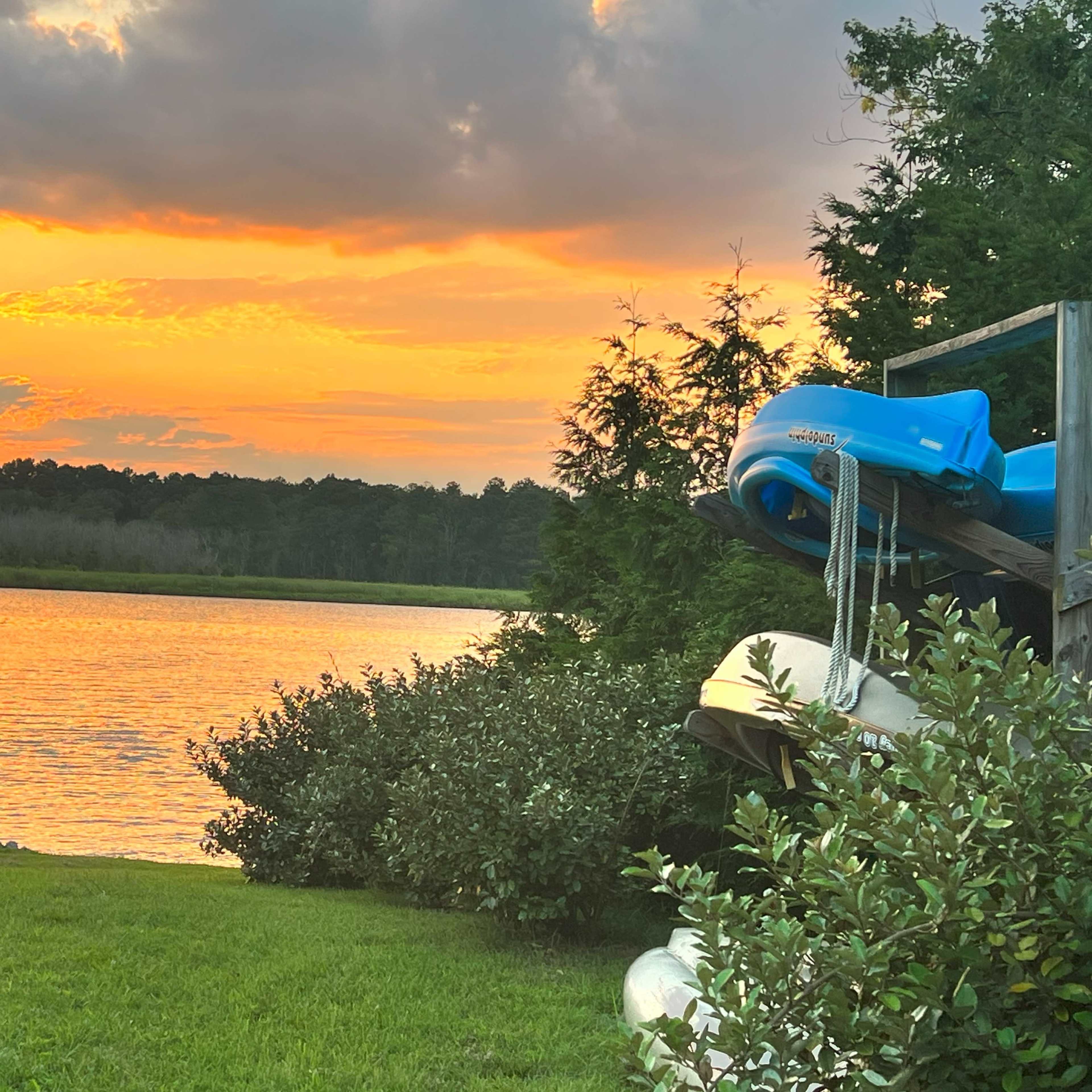 A serene lake at sunset with colorful reflections on the water, surrounded by greenery and kayaks stacked on a wooden structure.