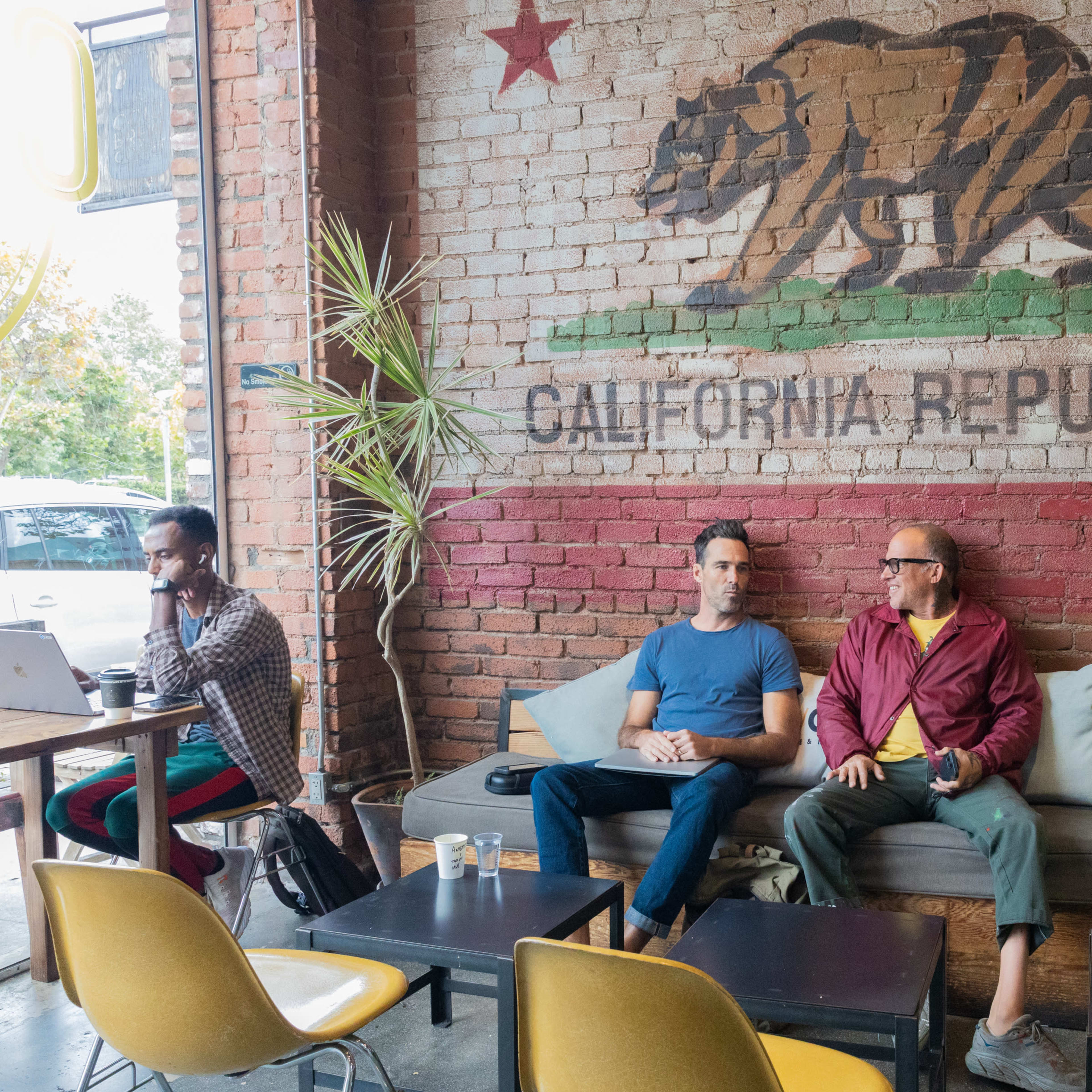 Three men are seated in a cafe with a California Republic mural on the brick wall behind them, while one man works on a laptop at a nearby table.