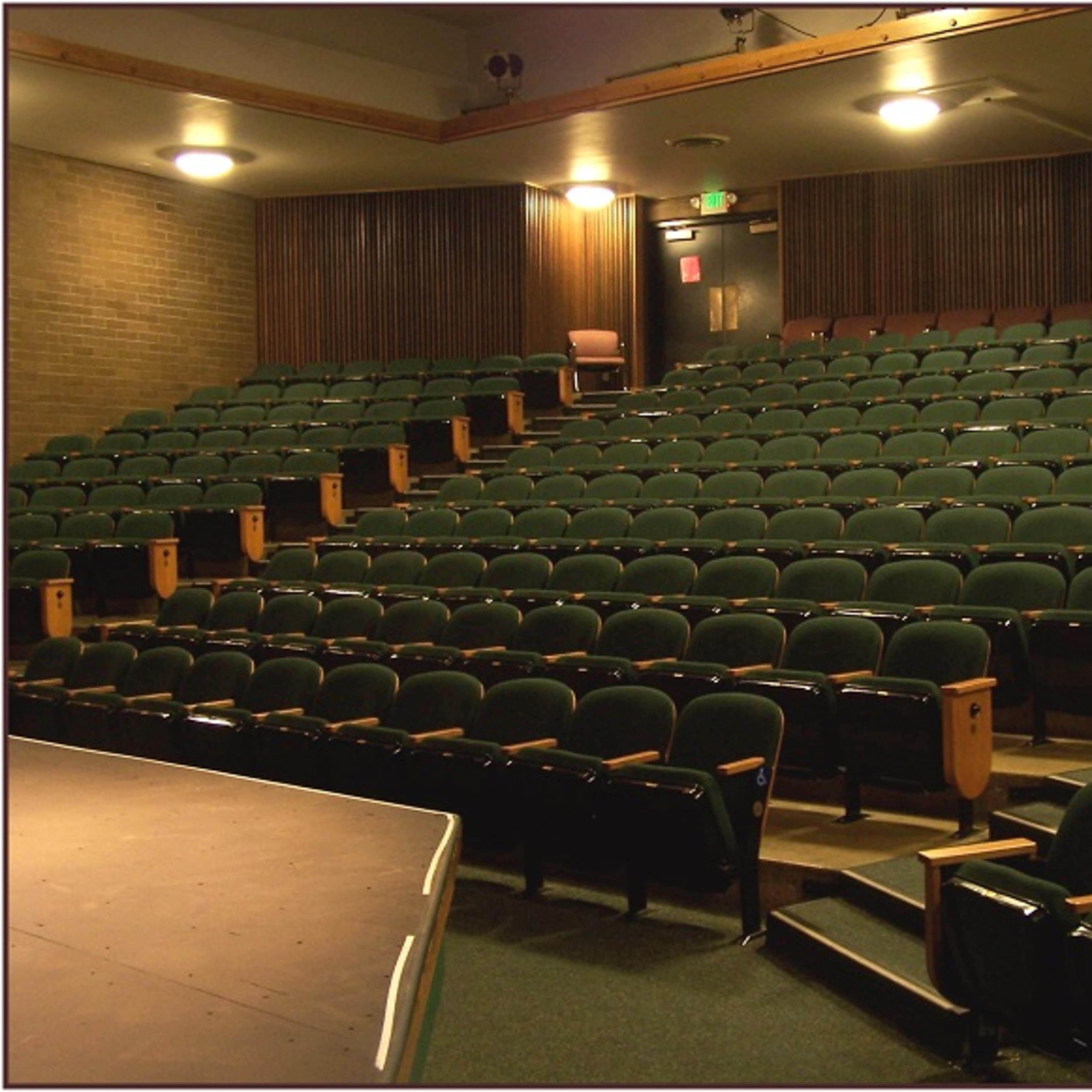 An empty theater with green upholstered seats arranged in rows facing a small elevated stage.