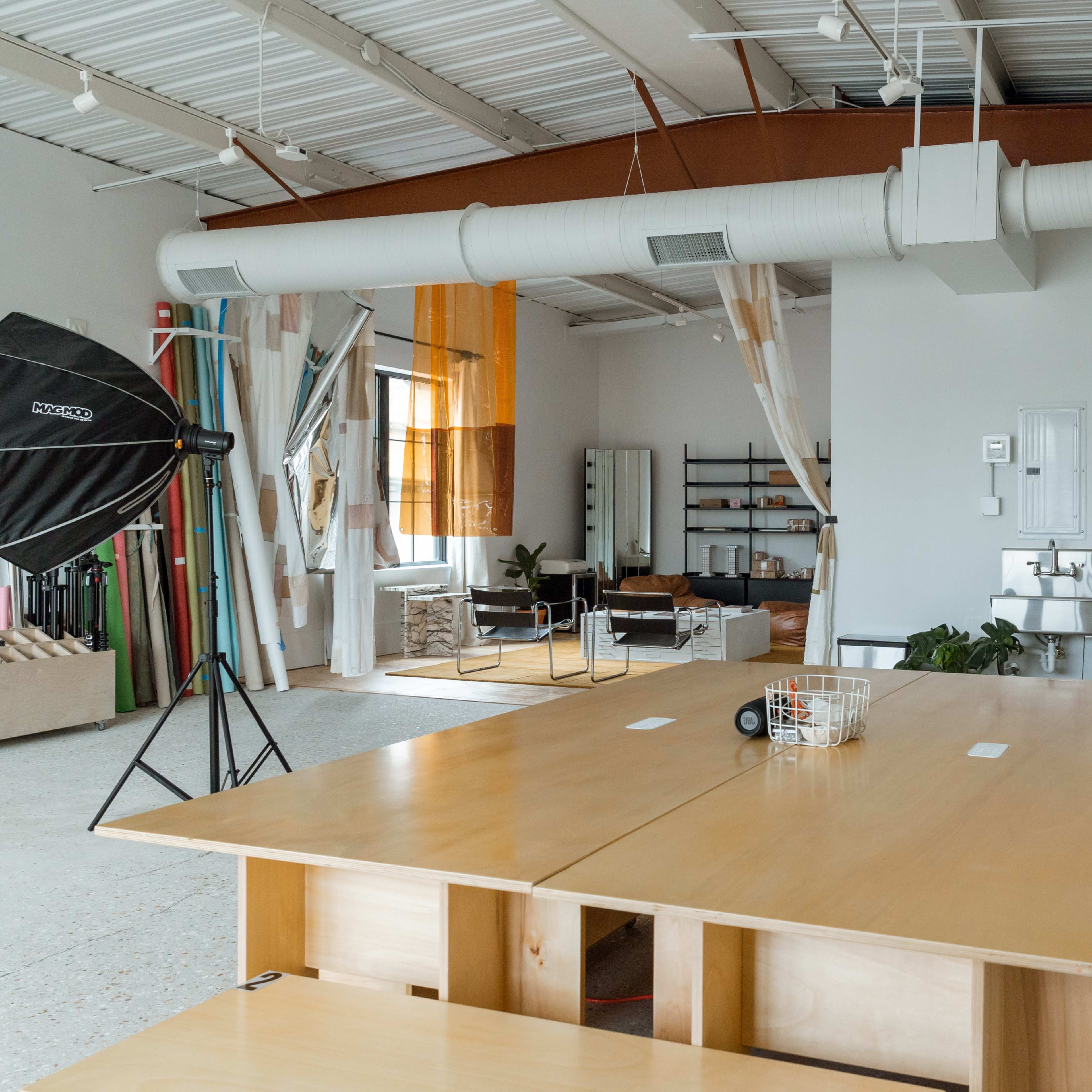 A spacious studio with a large wooden table, various lighting equipment, and a minimalist interior featuring shelves and couches.