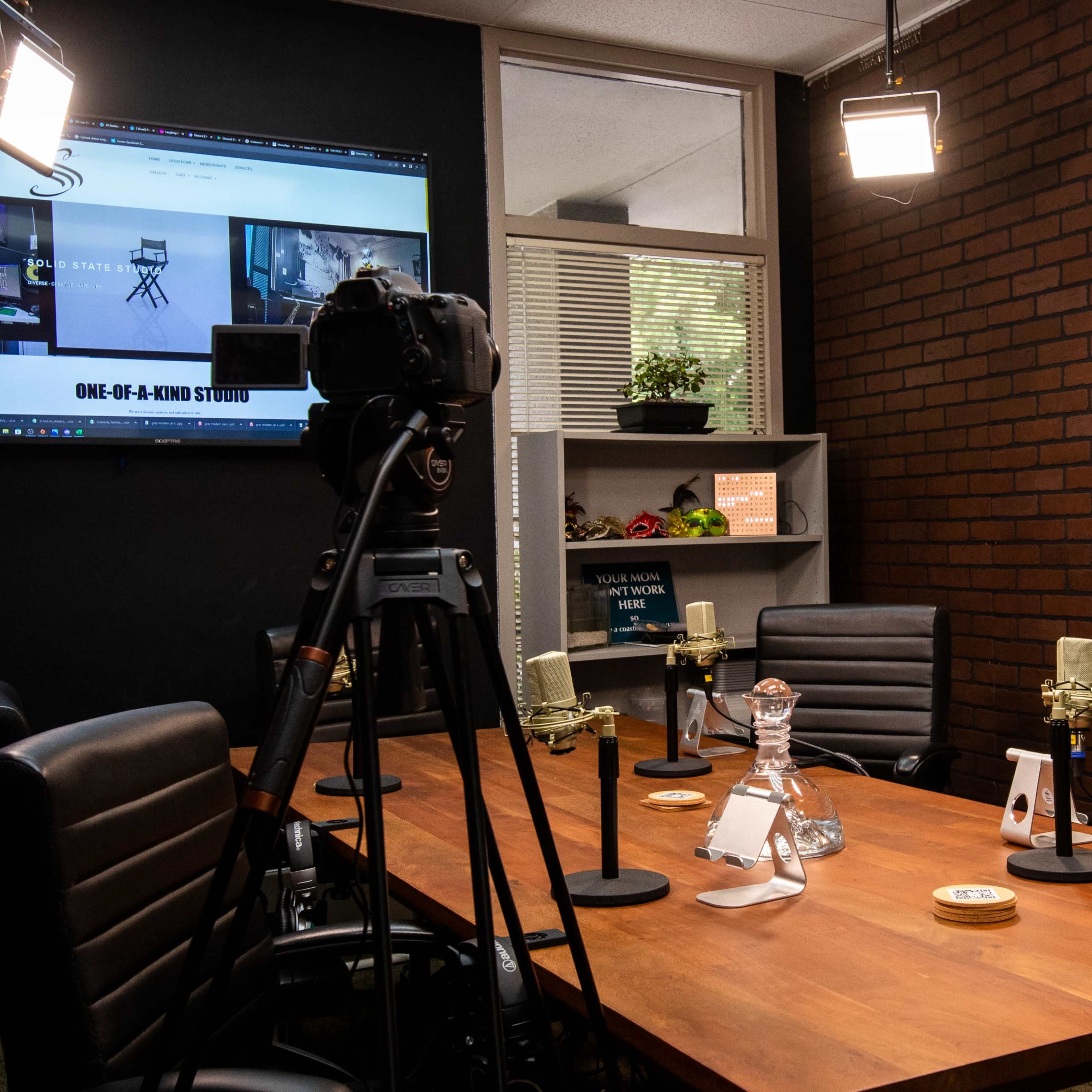 A video recording studio setup with a camera on a tripod, microphones on a wooden table, and a large screen displaying content on the wall.