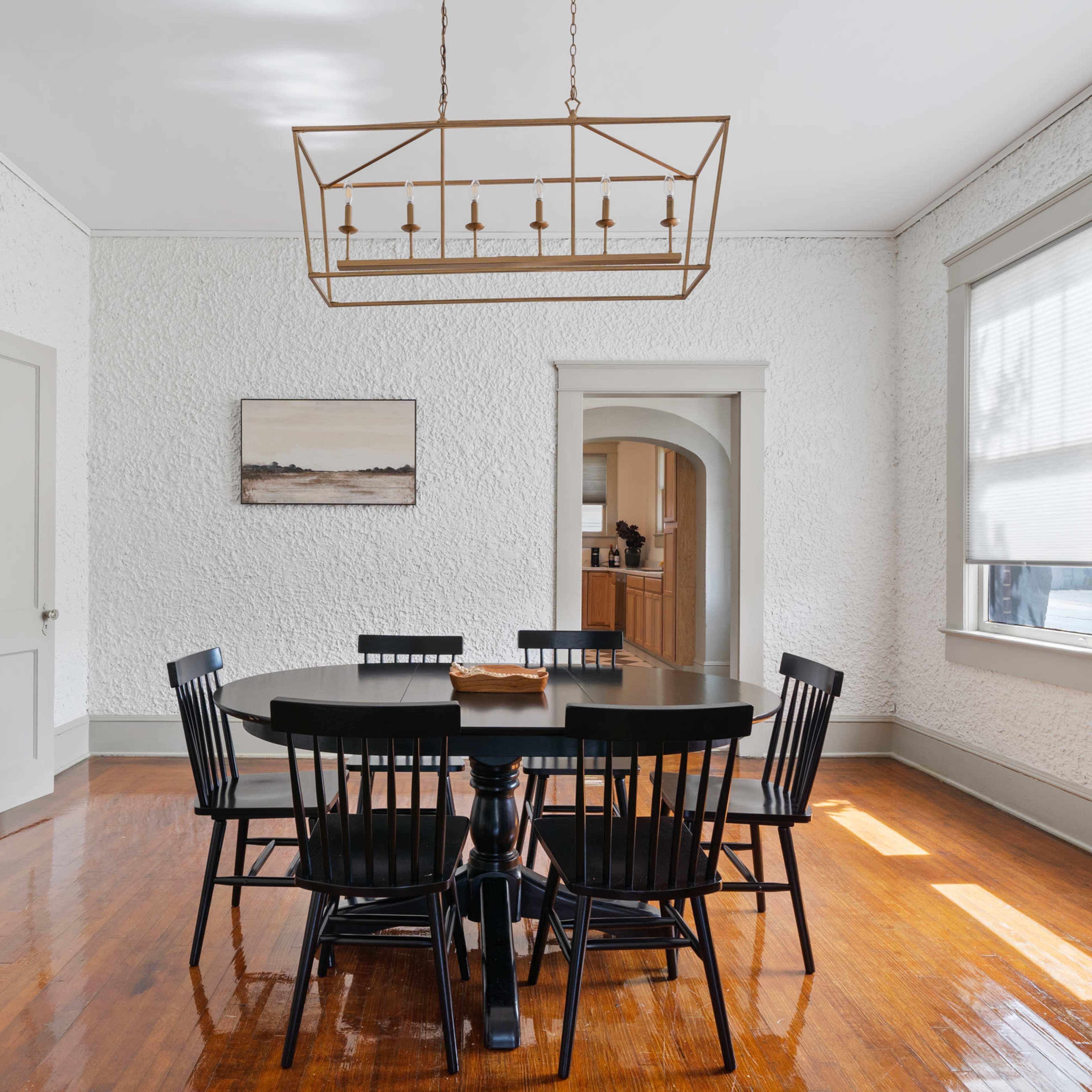 A round black dining table with matching chairs is positioned in a bright room with textured white walls and large windows.