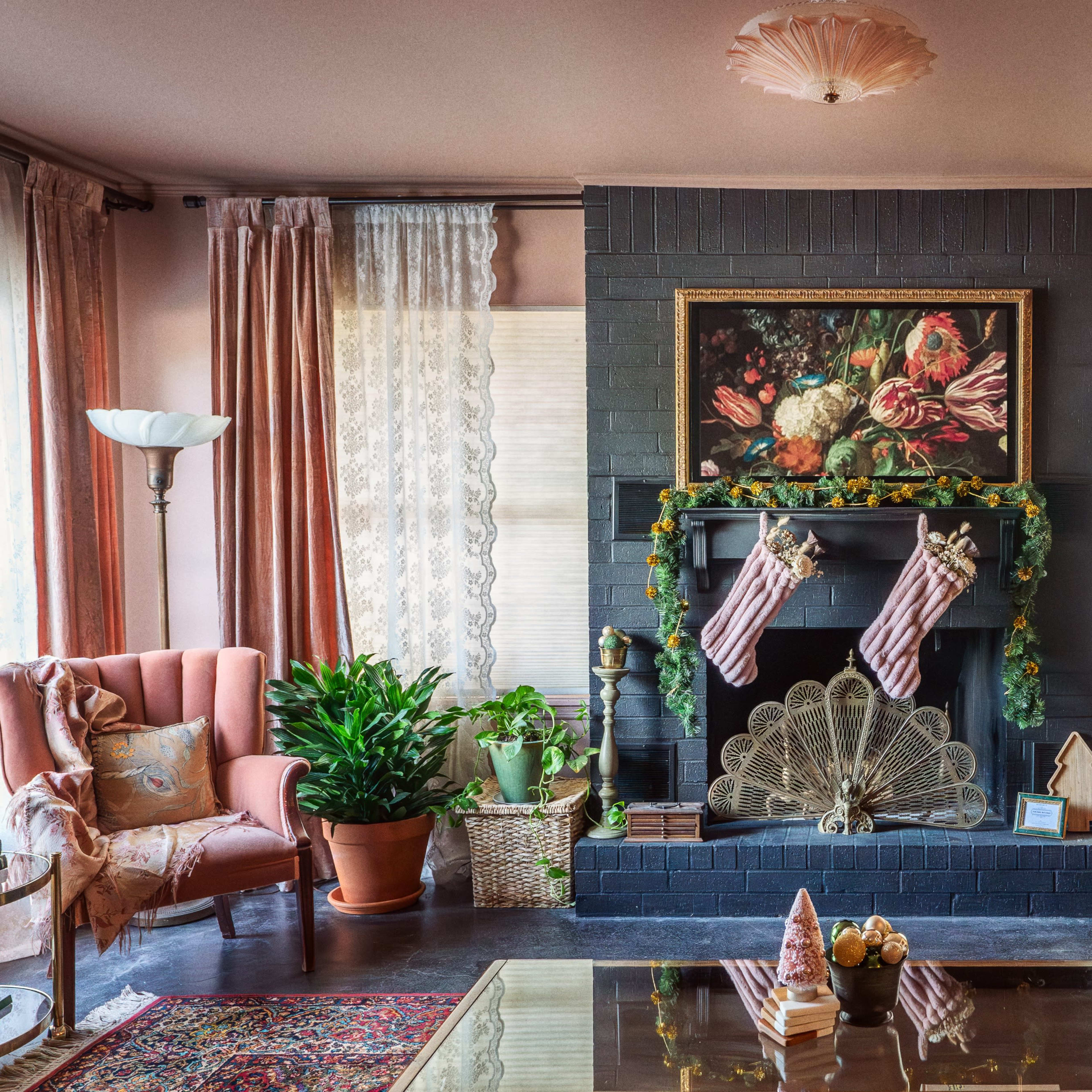 The living room features a fireplace adorned with stockings, a floral painting, and decorated with plants and furniture in warm colors.