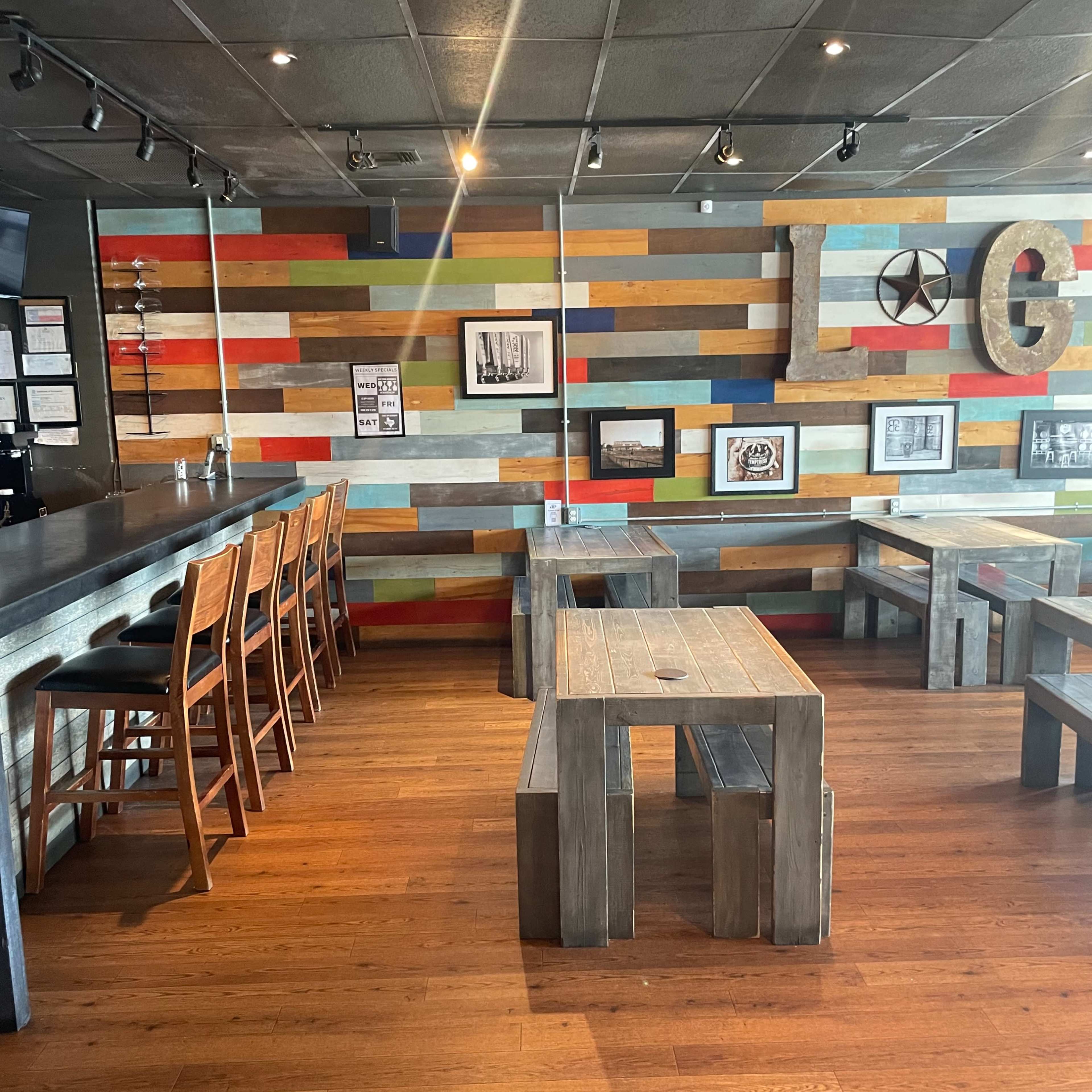 A restaurant interior featuring wooden benches and tables against a colorful, slatted wall, with a bar area on the left.