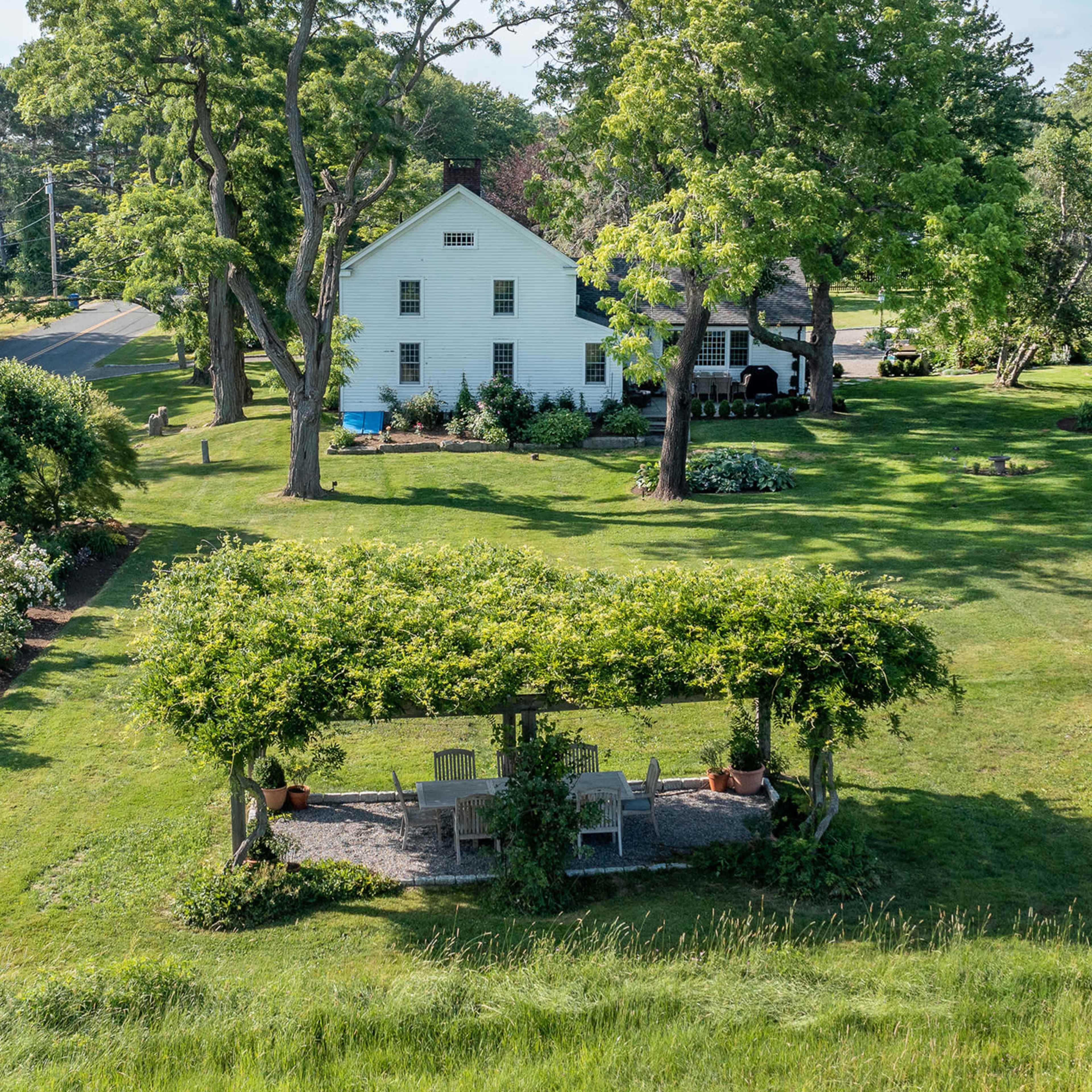 A spacious yard with a white house and a gazebo surrounded by trees and flowering plants.