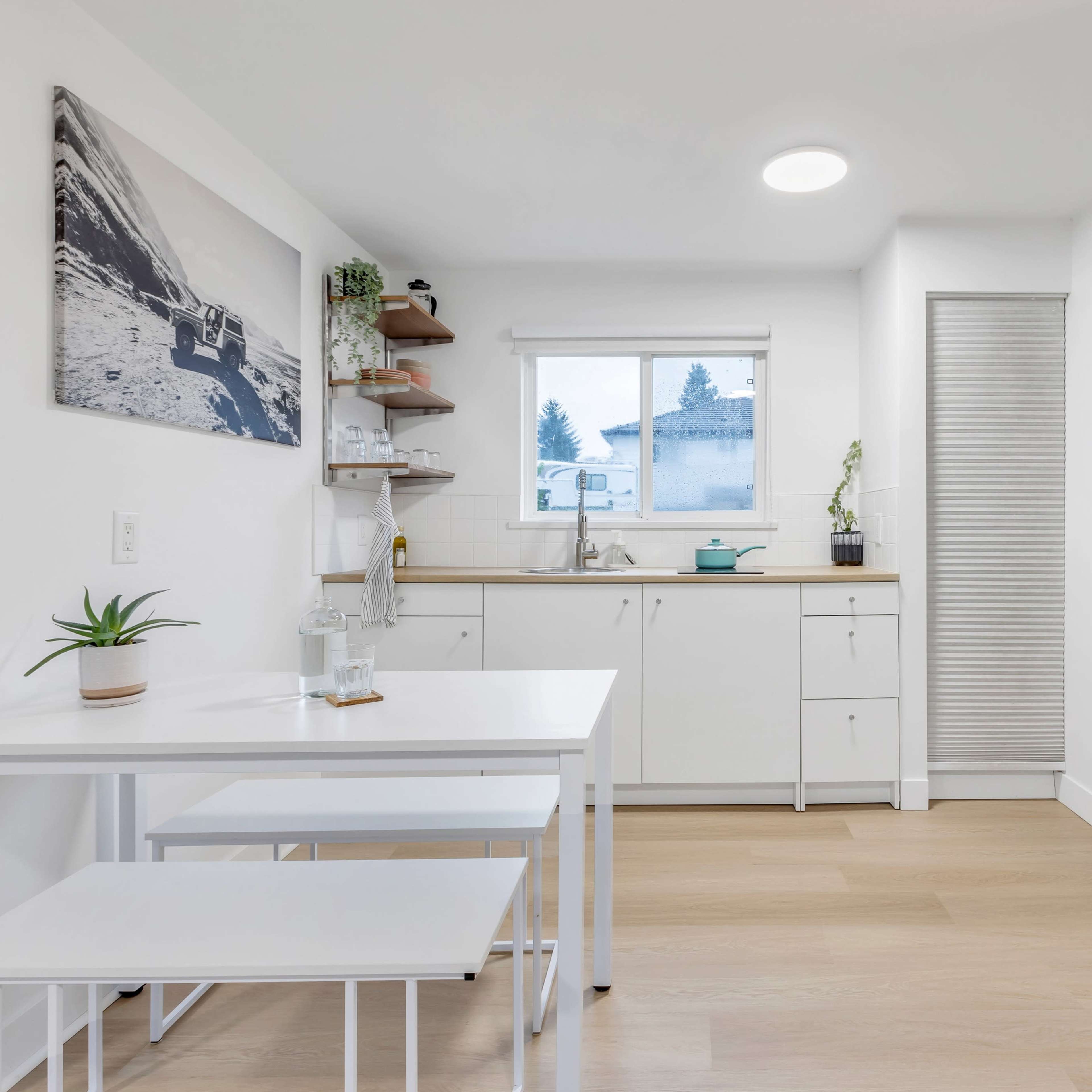 A modern kitchen features white cabinetry, a small dining table, and a refrigerator, with a window providing natural light.