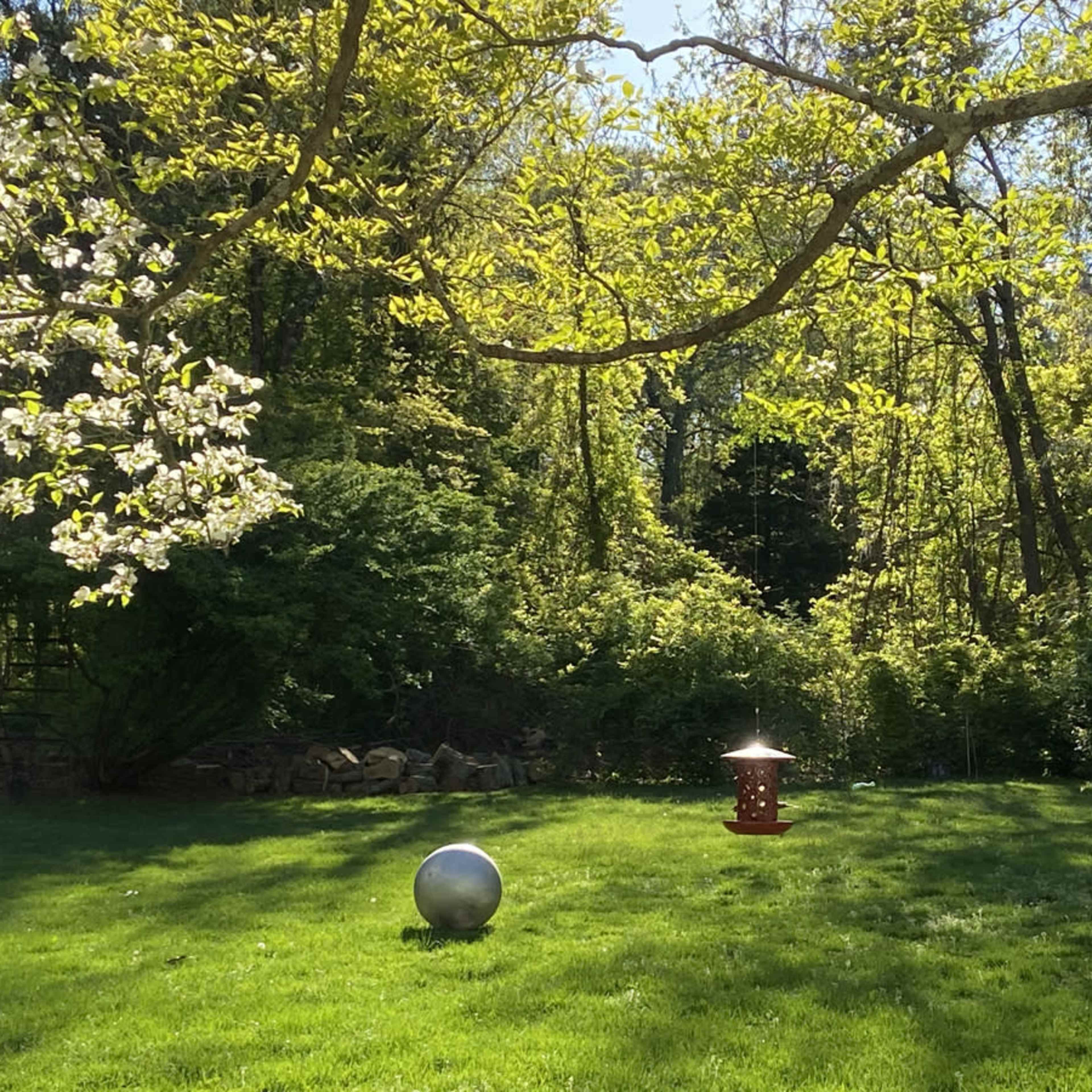 A spherical metal sculpture and a bird feeder are situated on a green lawn under a canopy of trees.