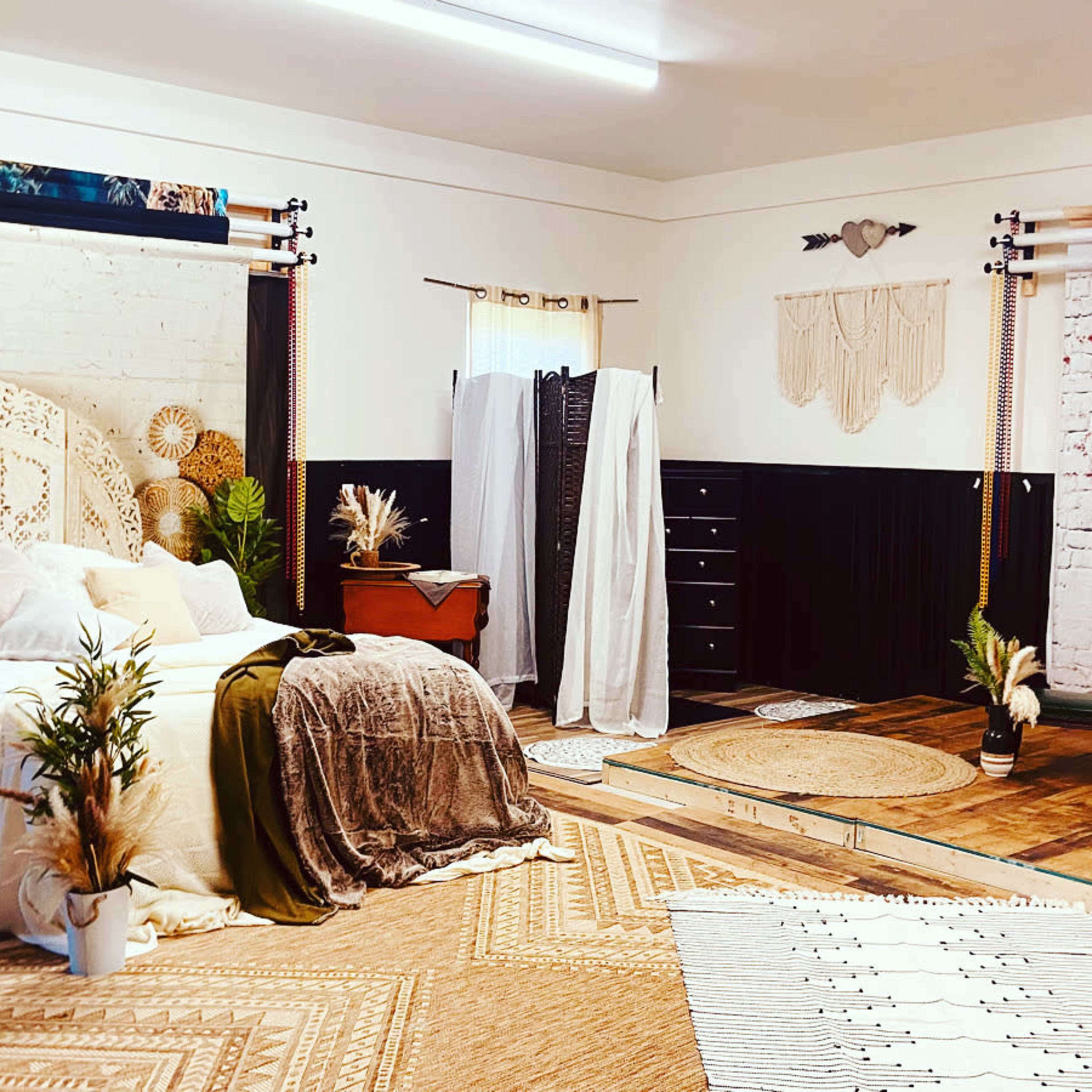 A styled interior room featuring a bed with woven headboard, various decorative plants, and distinct seating areas separated by patterned rugs and backdrops.