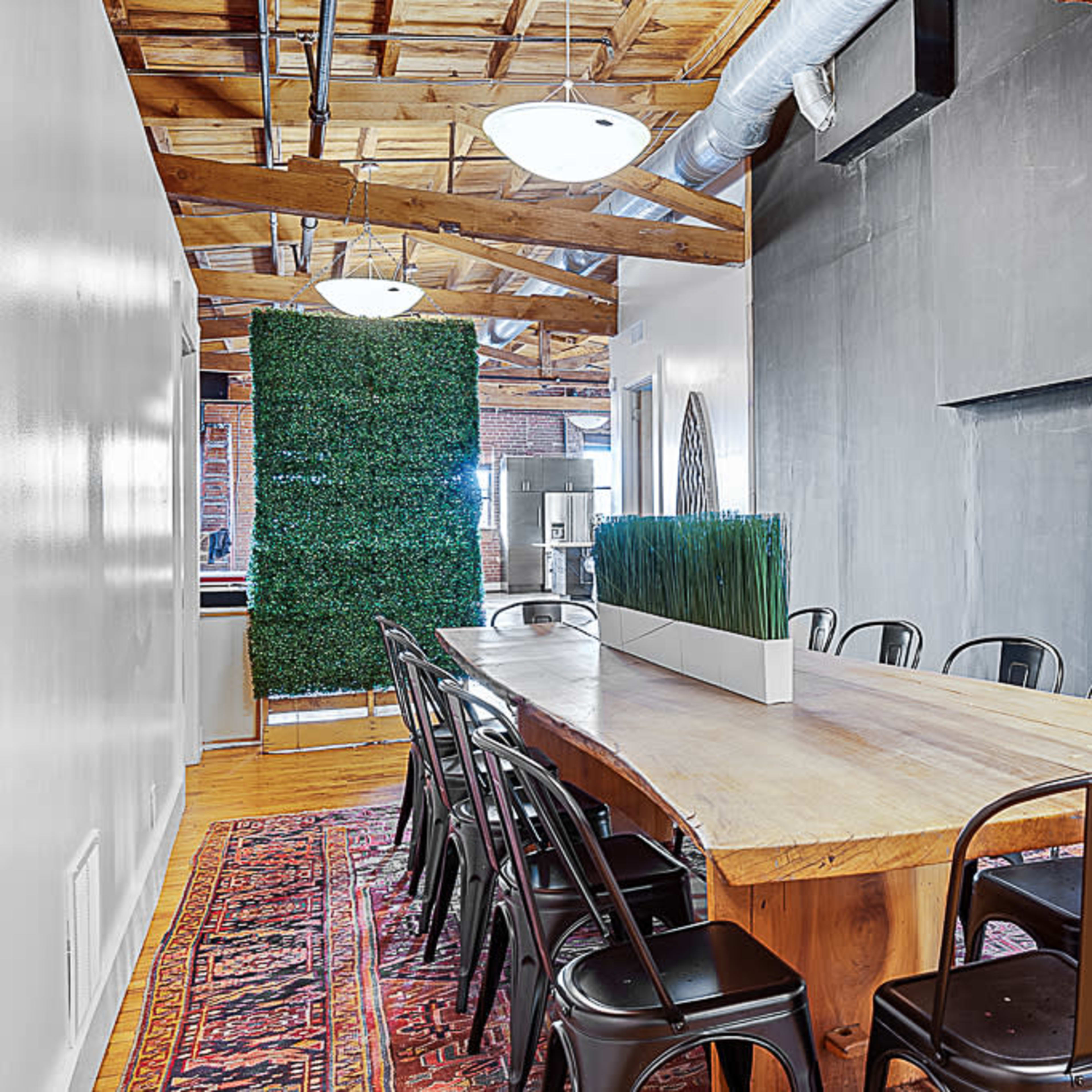A dining area featuring a large wooden table with black metal chairs, a green plant installation, and a chalkboard wall.