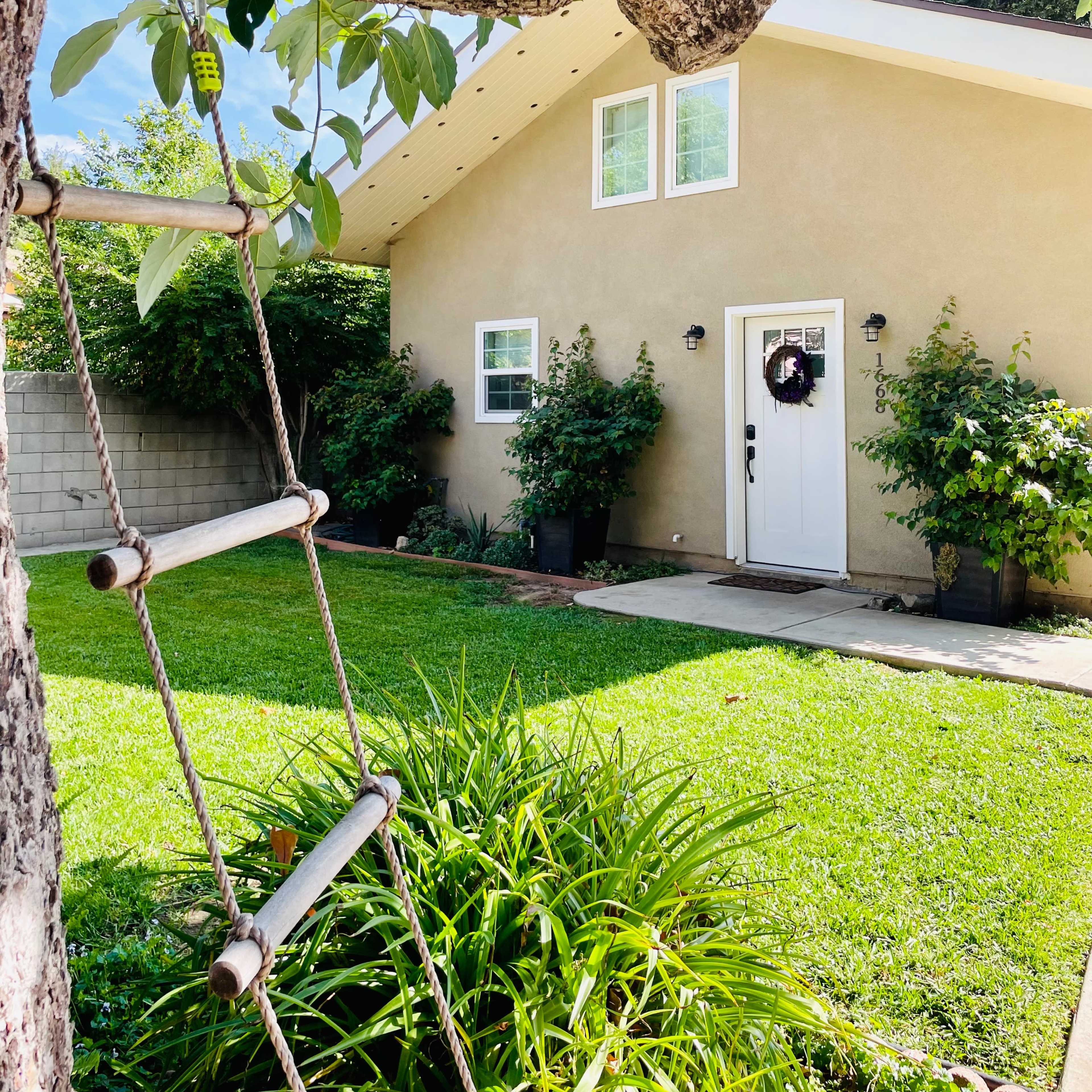 A tree with a rope ladder stands next to a house with a light-colored exterior and a front door adorned with a wreath, surrounded by greenery.