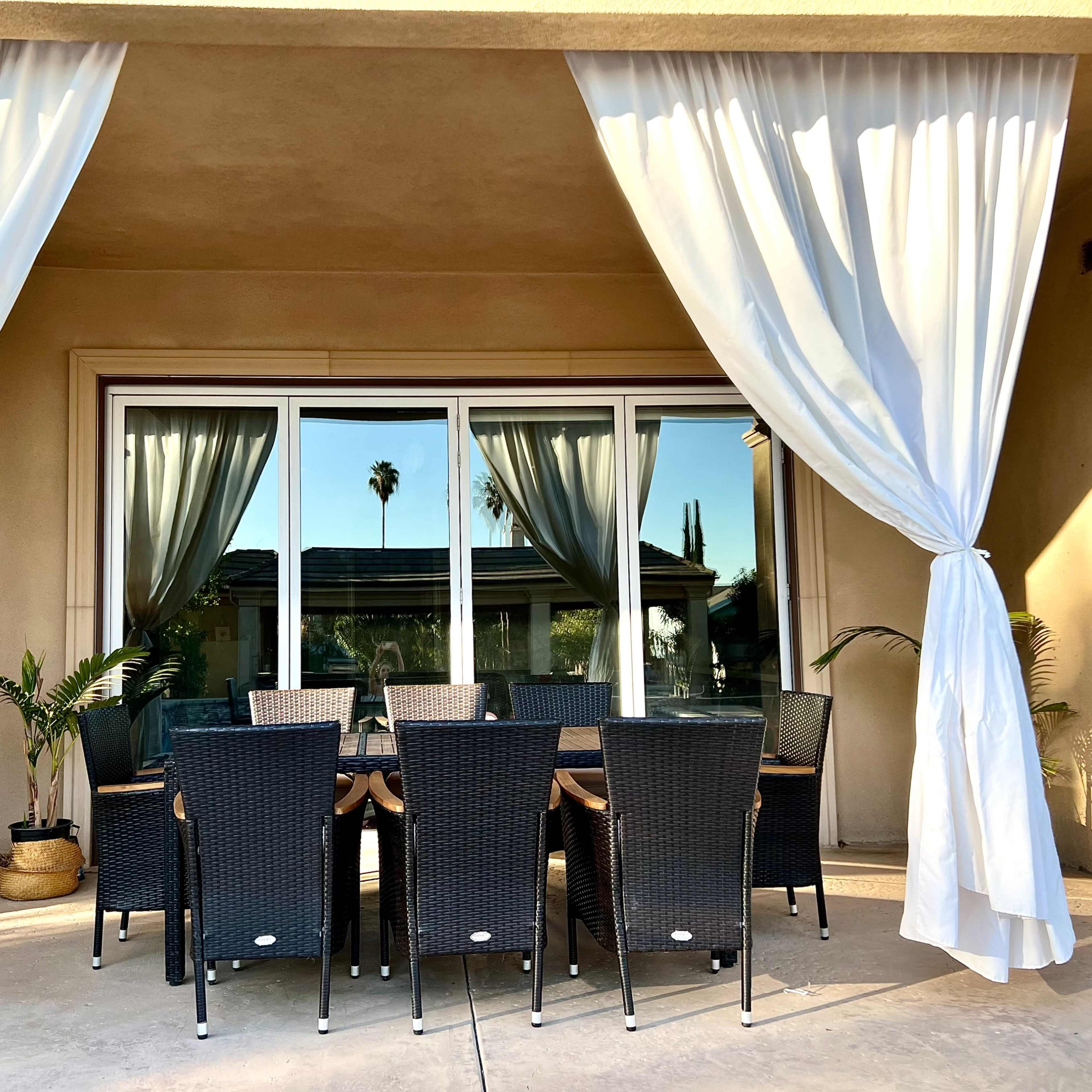 A dining table with chairs is set under a canopy of white curtains, with palm trees reflected in the large window behind it.