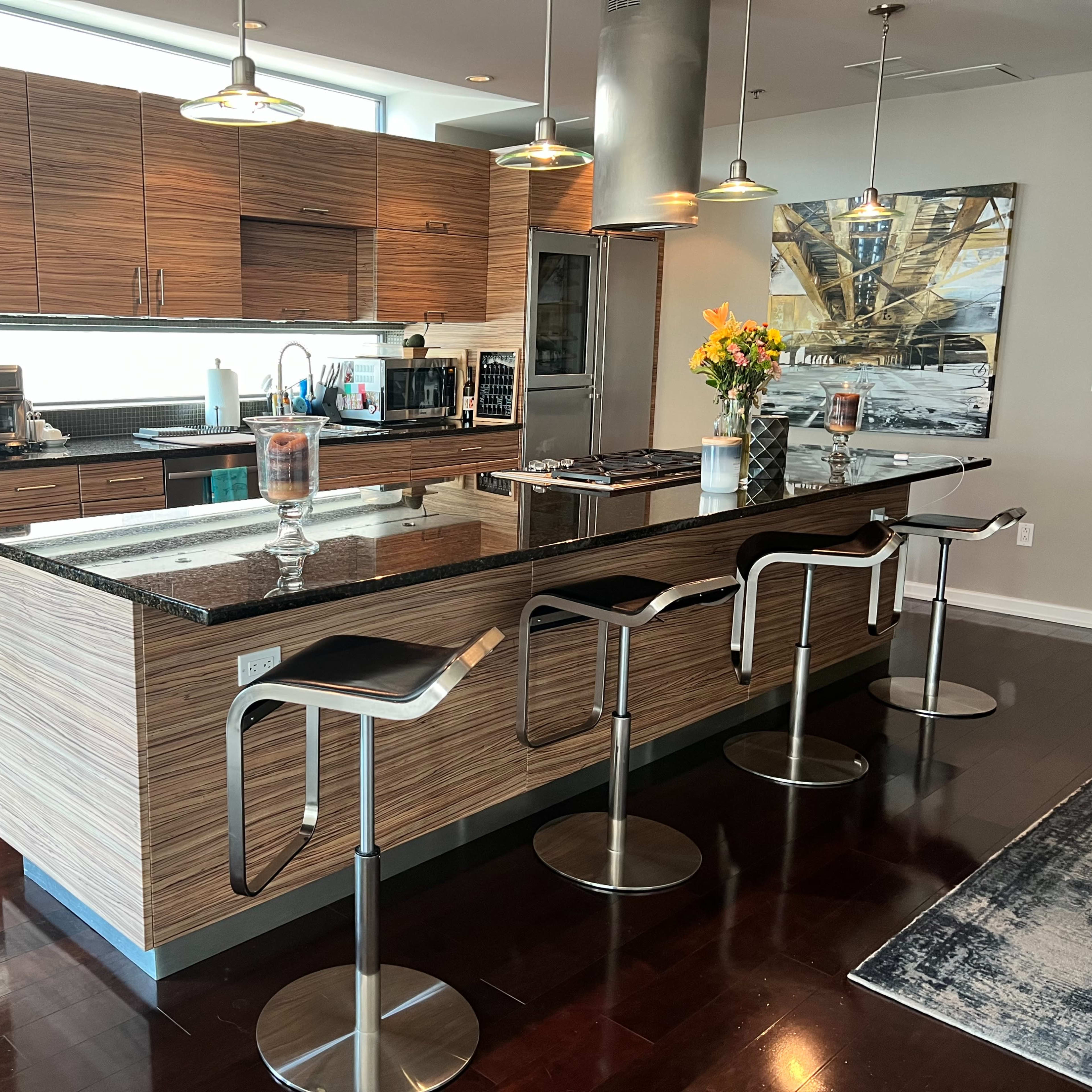 A modern kitchen with a central island, bar stools, and wooden cabinetry, featuring appliances and a vase of flowers.