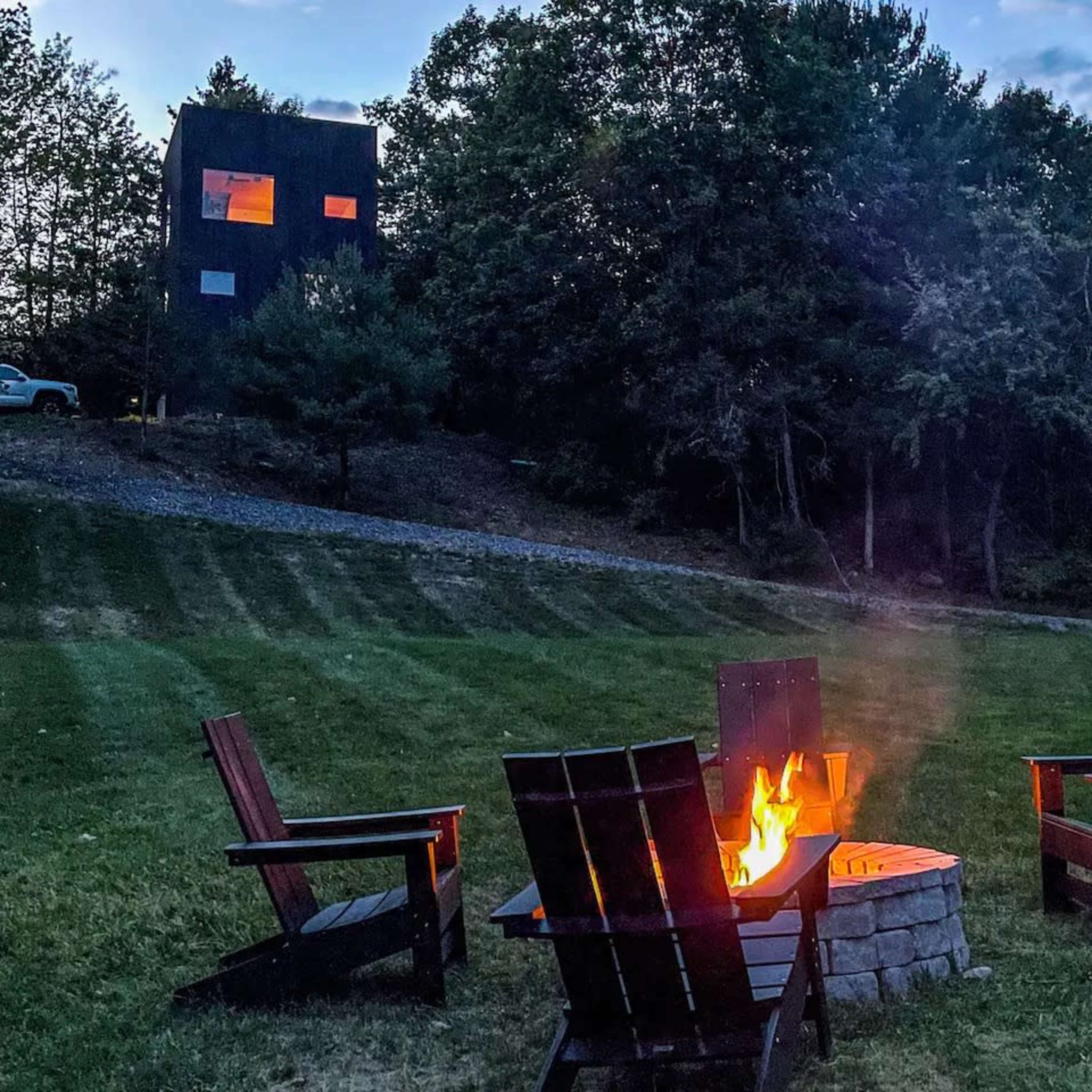 A fire pit with wooden chairs is set up in a grassy area, facing a modern, dark-colored building nestled among trees.