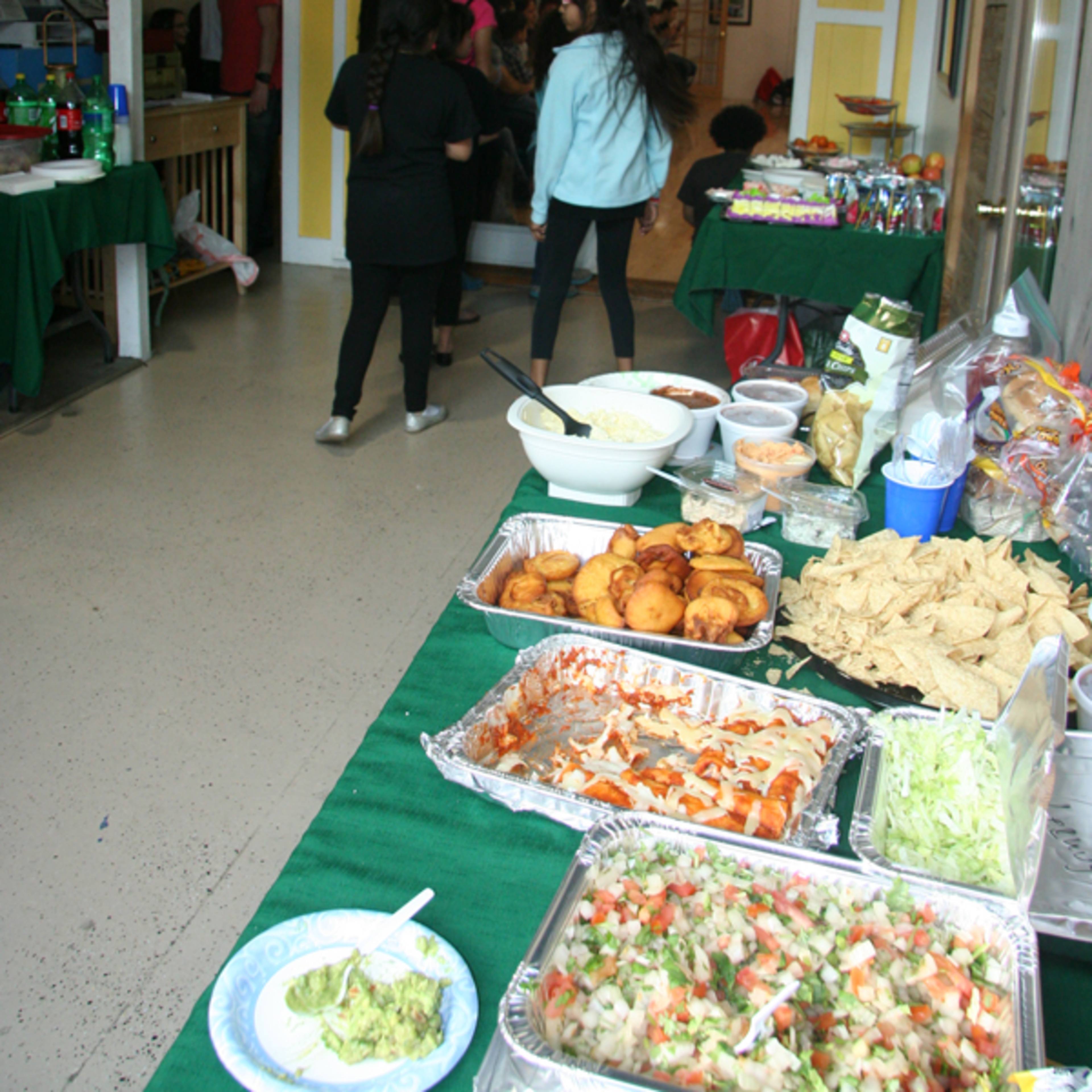 A gathering space with tables covered in green cloths, displaying various prepared dishes and snacks, while people are walking toward a doorway in the background.