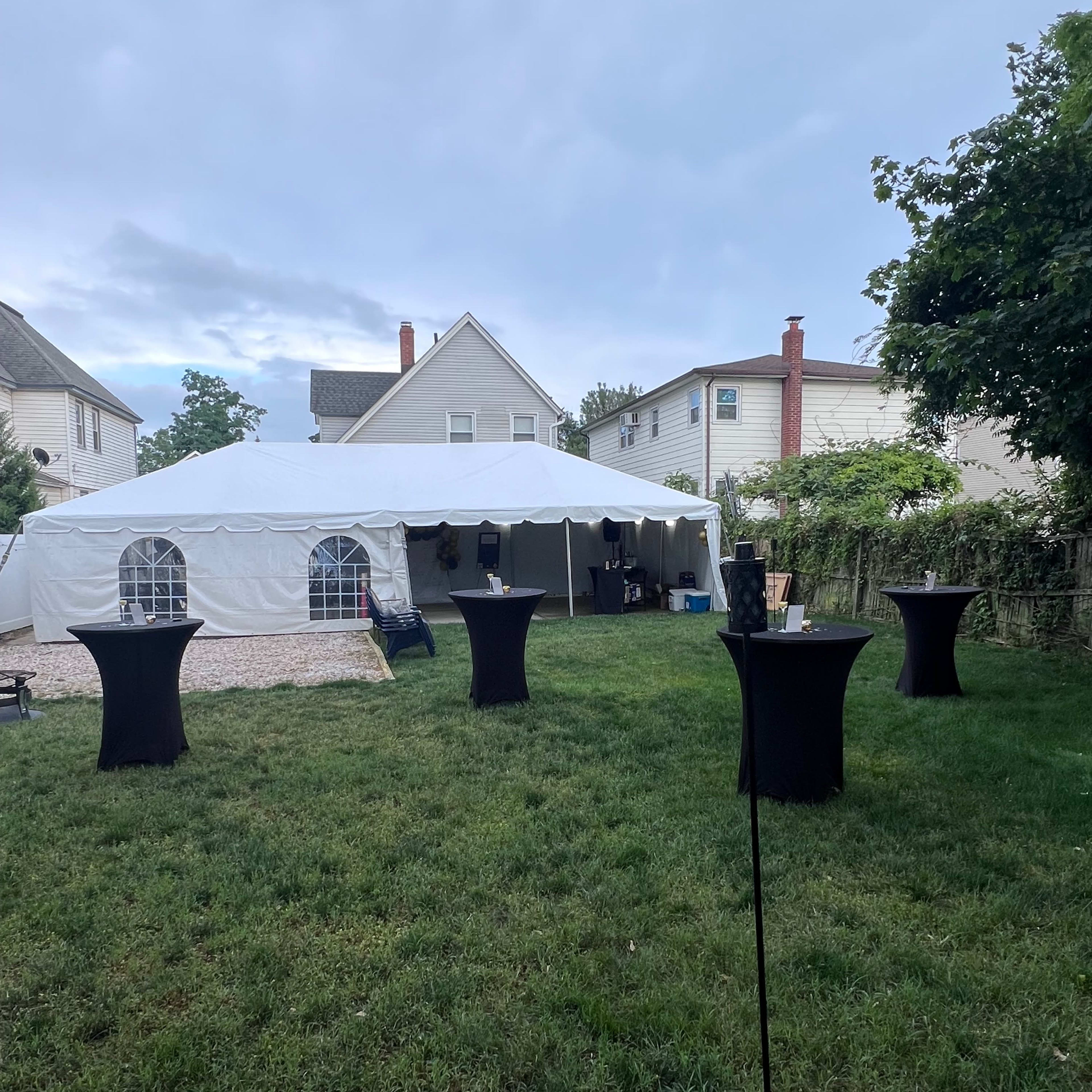 A white canopy tent is set up in a grassy backyard, surrounded by tall tables.