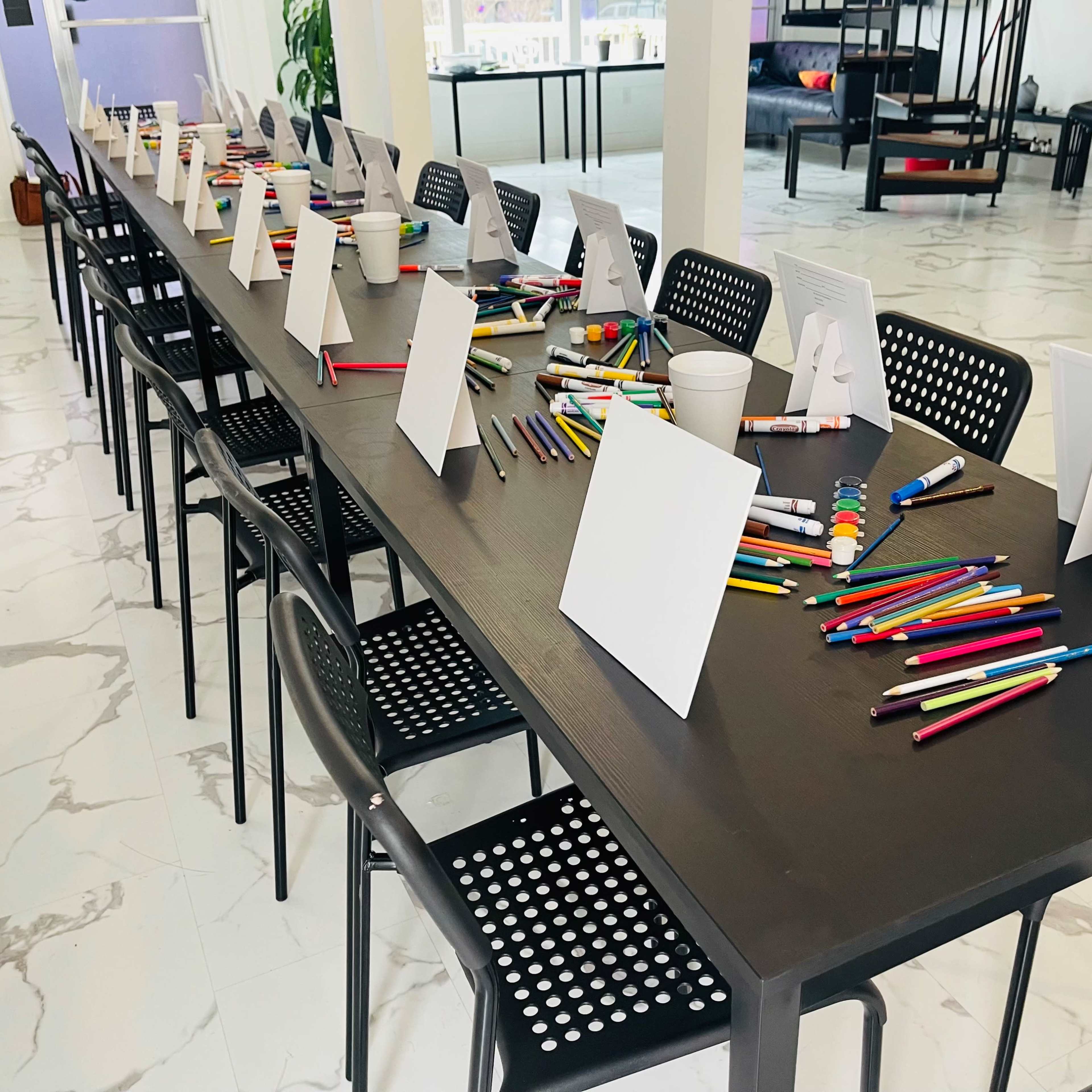 A long table is set up with various colored pens and blank cards, ready for an activity, surrounded by black chairs in a brightly lit room.