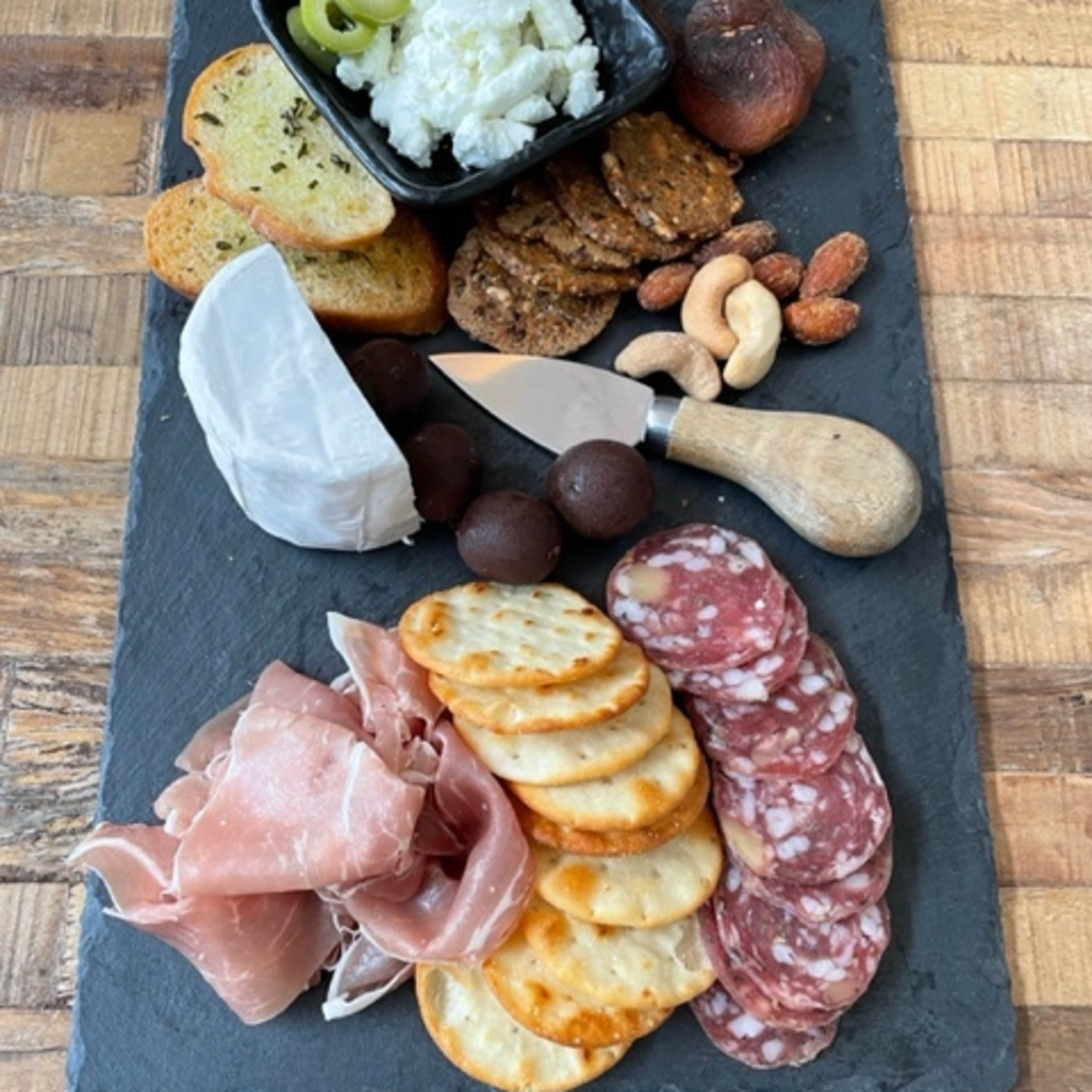 A black slate platter displays an assortment of crackers, cured meats, cheese, nuts, olives, and a small bowl of spreadable cheese.