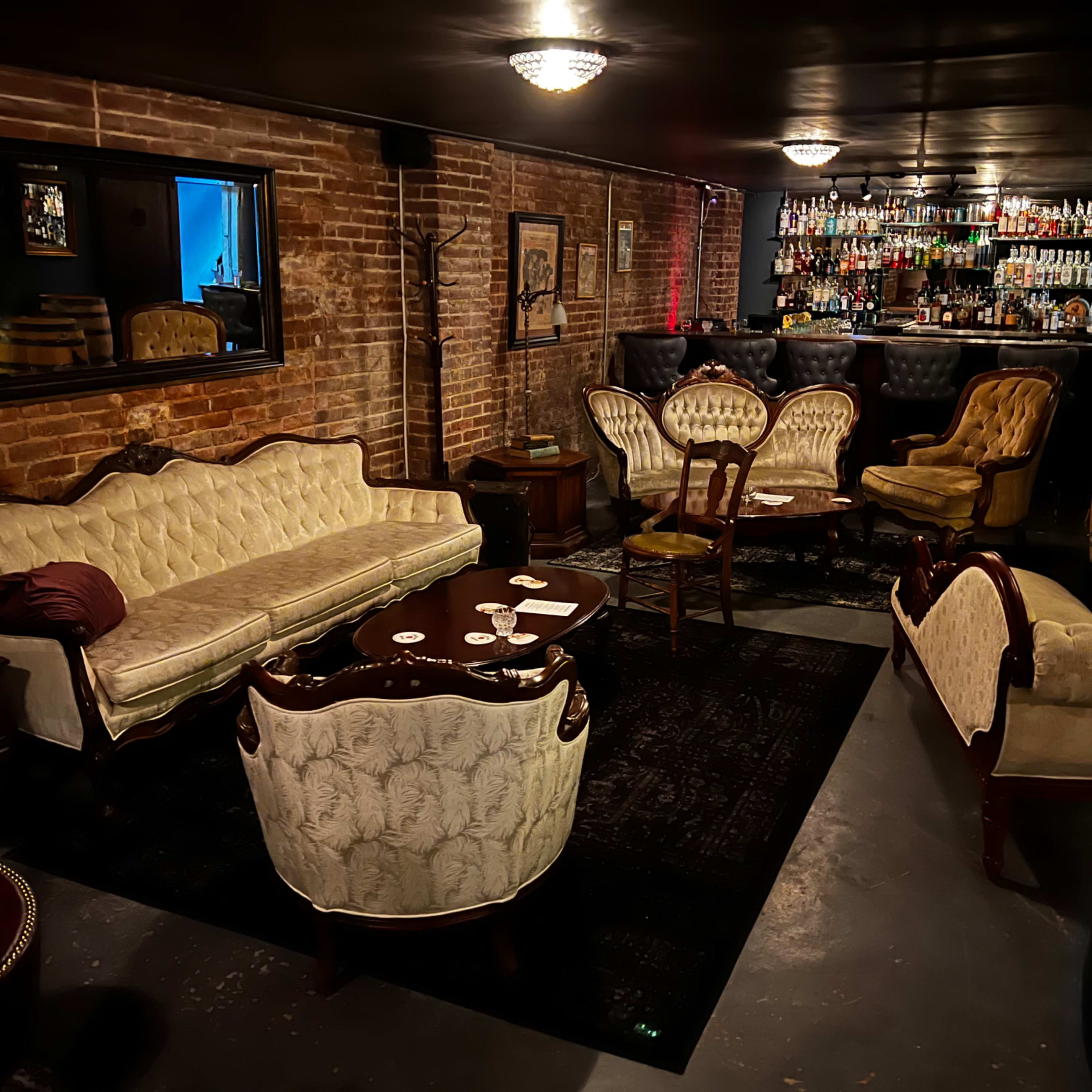 A dimly lit lounge with vintage-style furniture, including ornate sofas and chairs, arranged around a round table in front of a bar lined with liquor bottles.