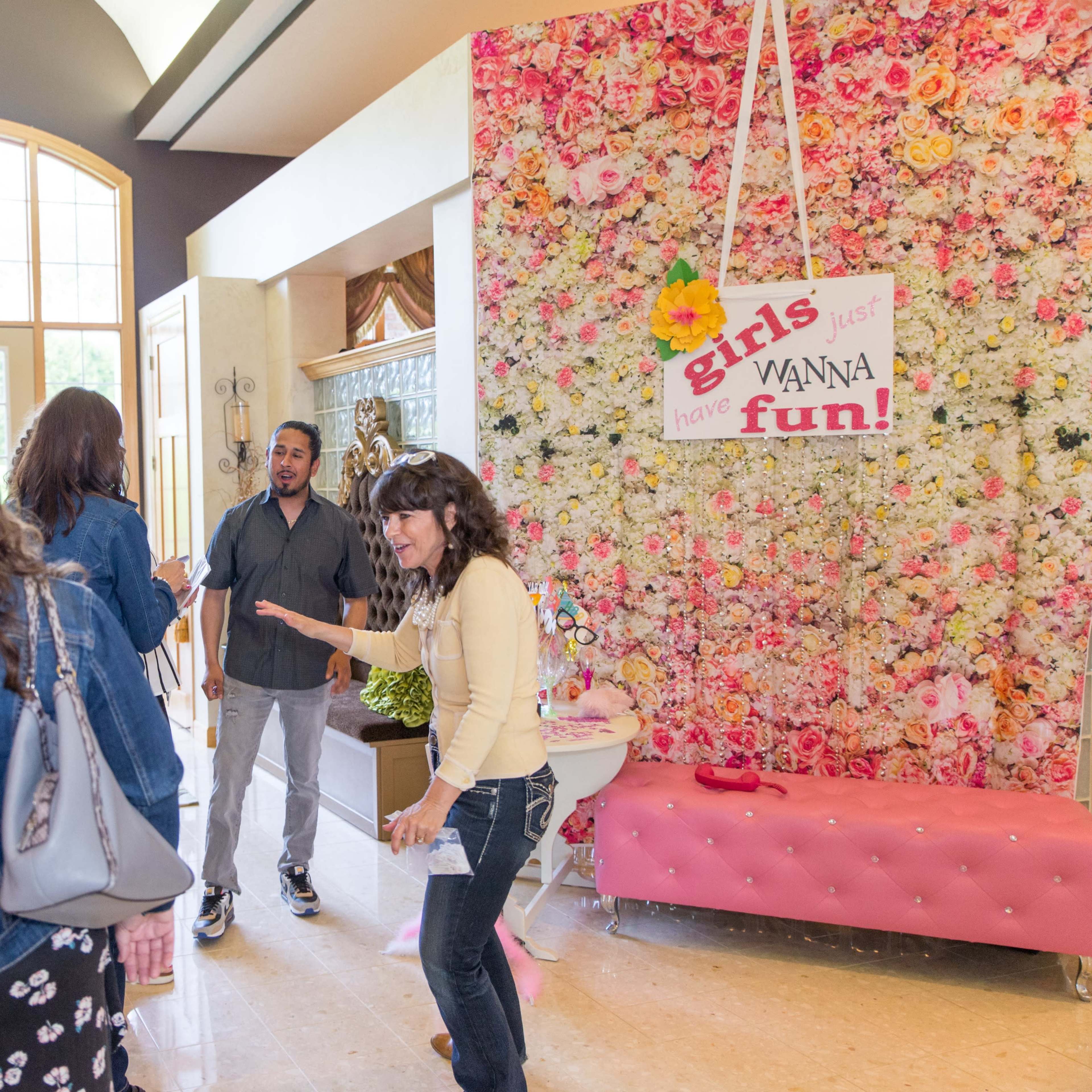 A group of people mingle in a lobby adorned with a vibrant floral backdrop and a sign that reads "girls just wanna have fun!"