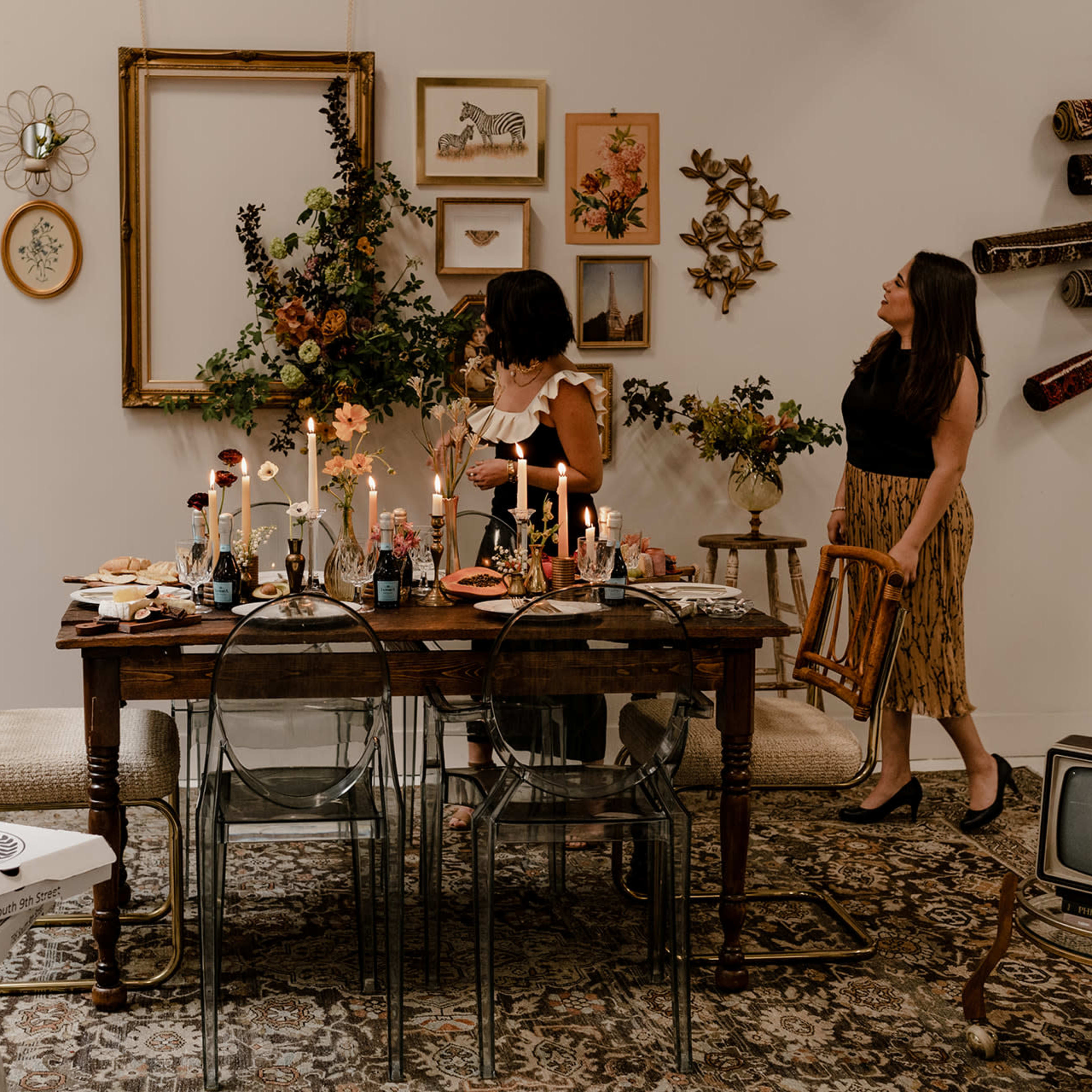 Two women stand beside a decorated dining table filled with food and candles, set against a backdrop of framed art and floral arrangements.