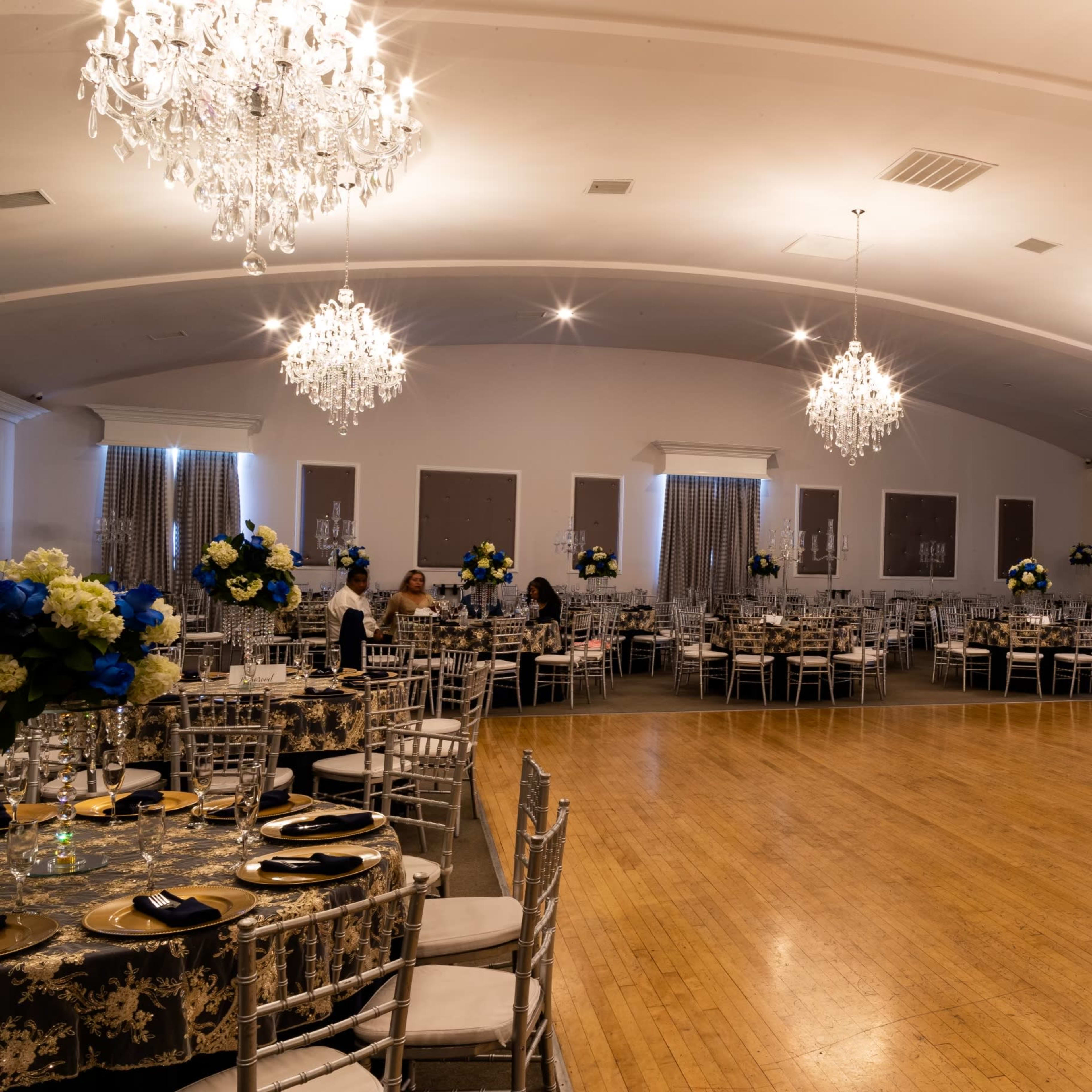 An elegantly decorated banquet hall with a wooden floor, round tables set with floral arrangements, and multiple chandeliers hanging from the ceiling.