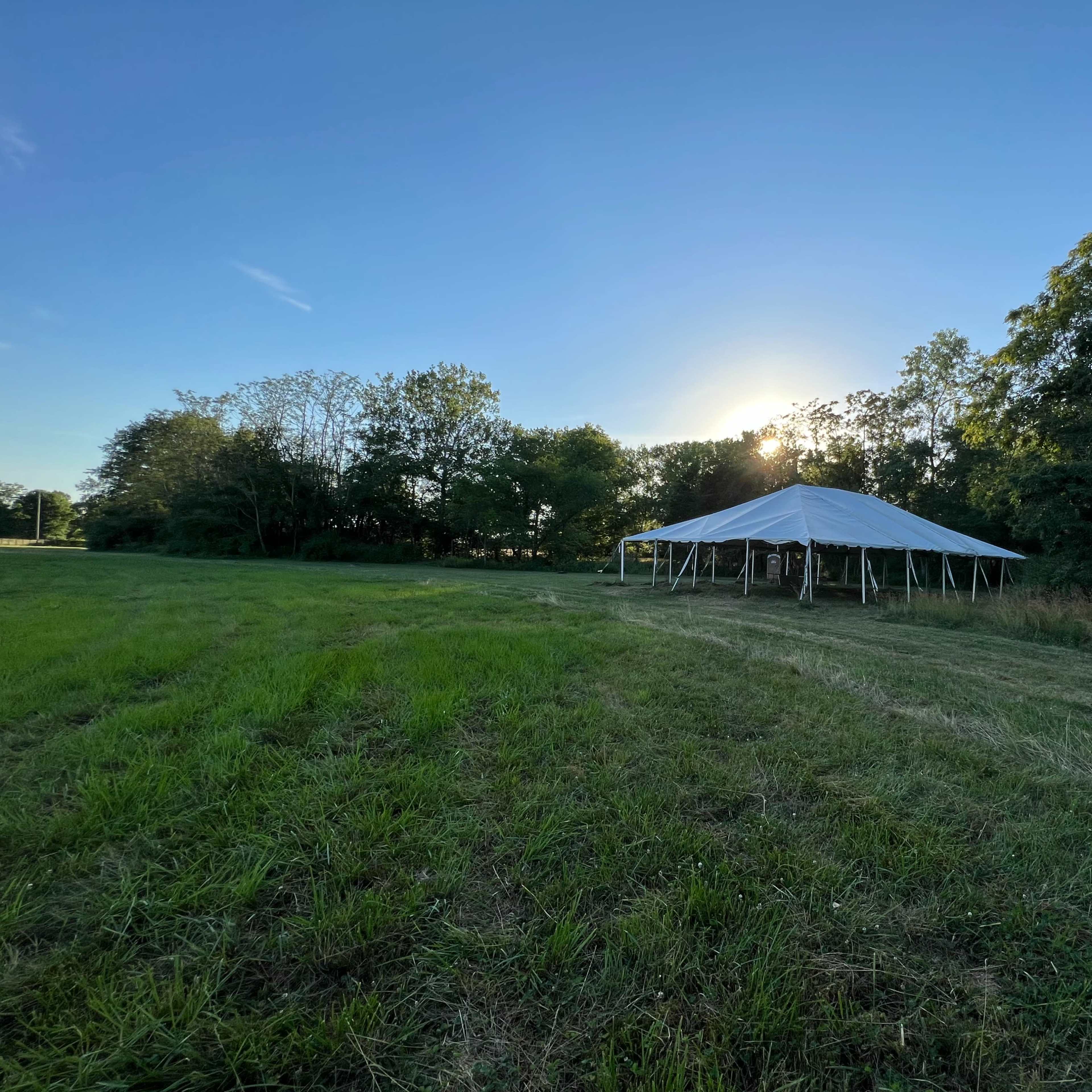 A white tent is set up on a grassy field with trees in the background and the sun illuminating the scene.